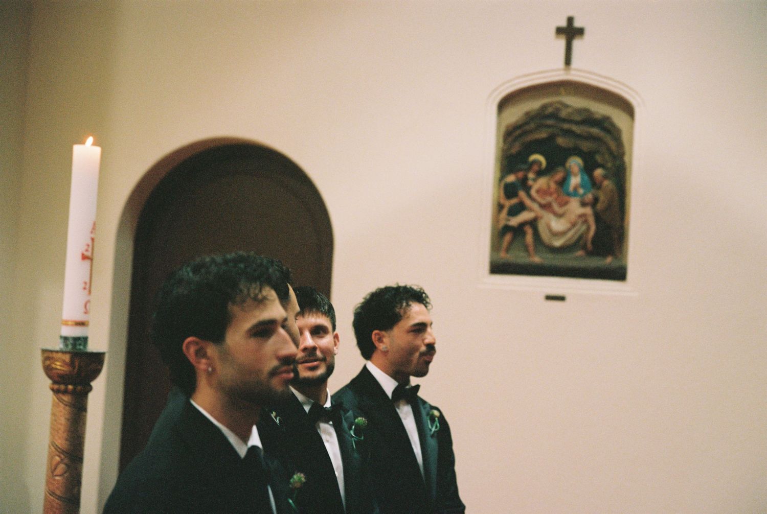 Three groomsmen standing at church altar during wedding ceremony with candle and religious artwork.