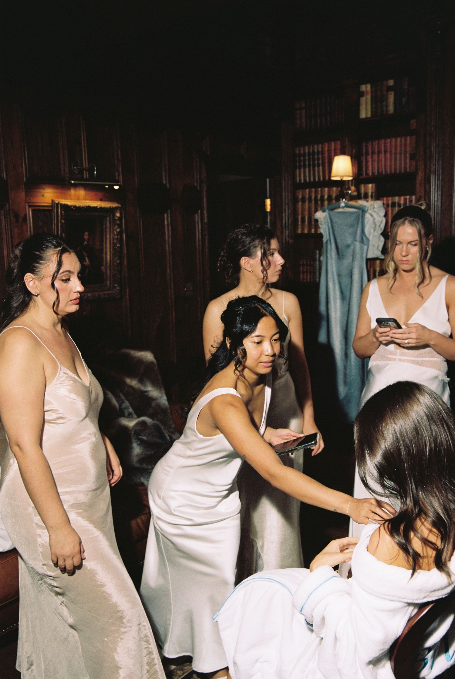 Bridesmaids in satin slip dresses getting ready together in dark wood-paneled room before wedding ceremony.