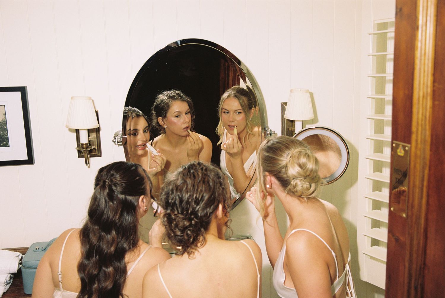 Bridesmaids in satin slips touch up makeup together in a round mirror.