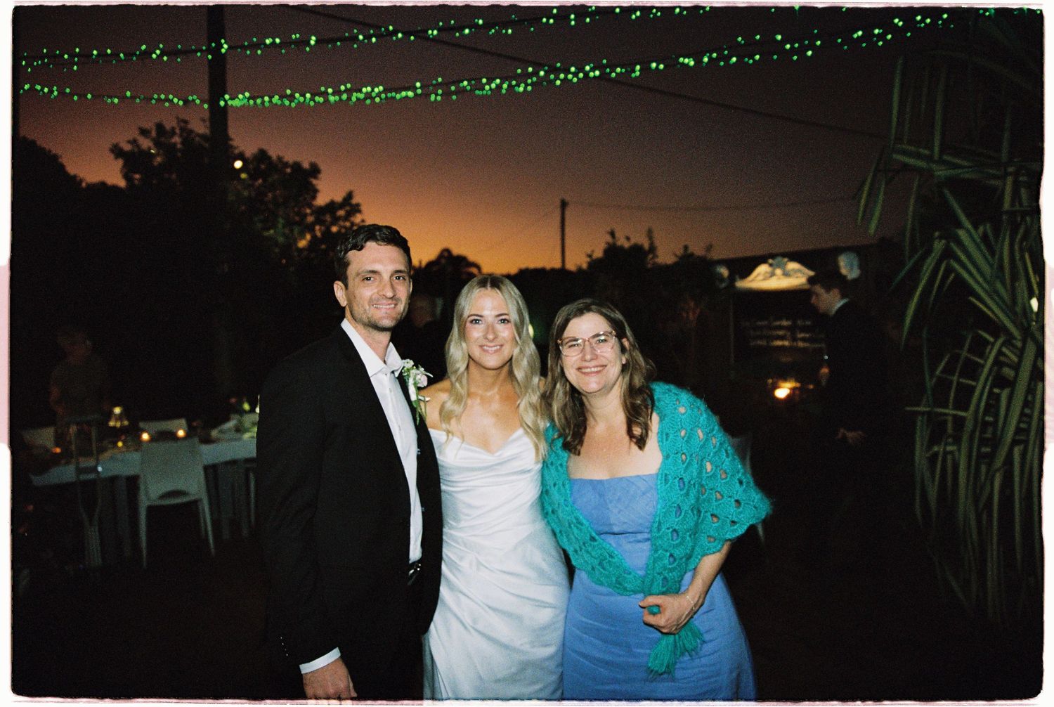 Three wedding guests pose outdoors at sunset with string lights overhead at an evening reception.