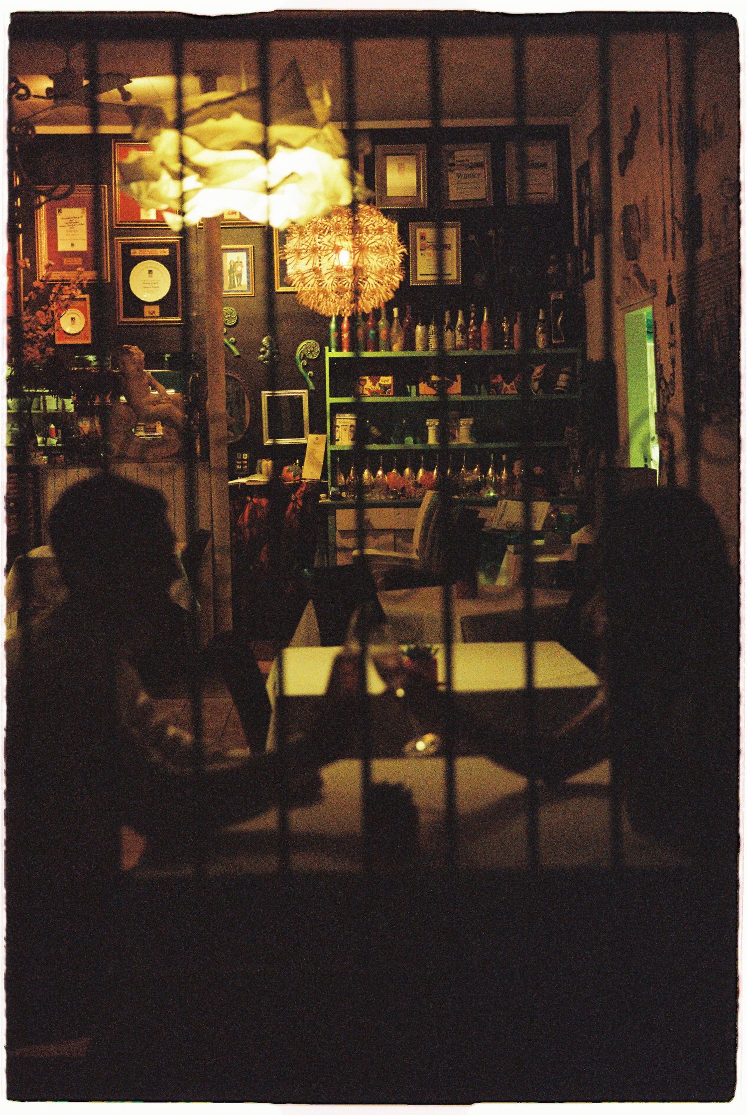 Silhouetted guests relax inside a warmly lit eclectic bar or restaurant viewed through iron bars.