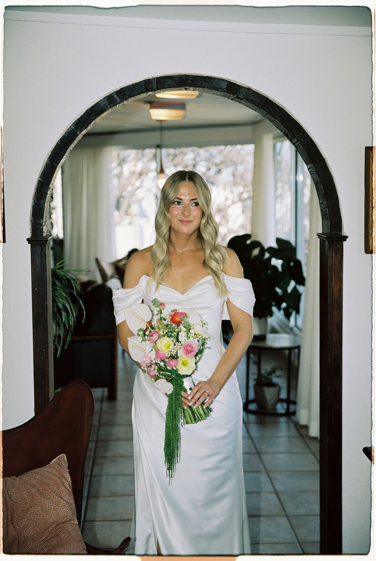 Bride in white off-shoulder gown holding pink bouquet stands under dark wood arch in elegant indoor setting.