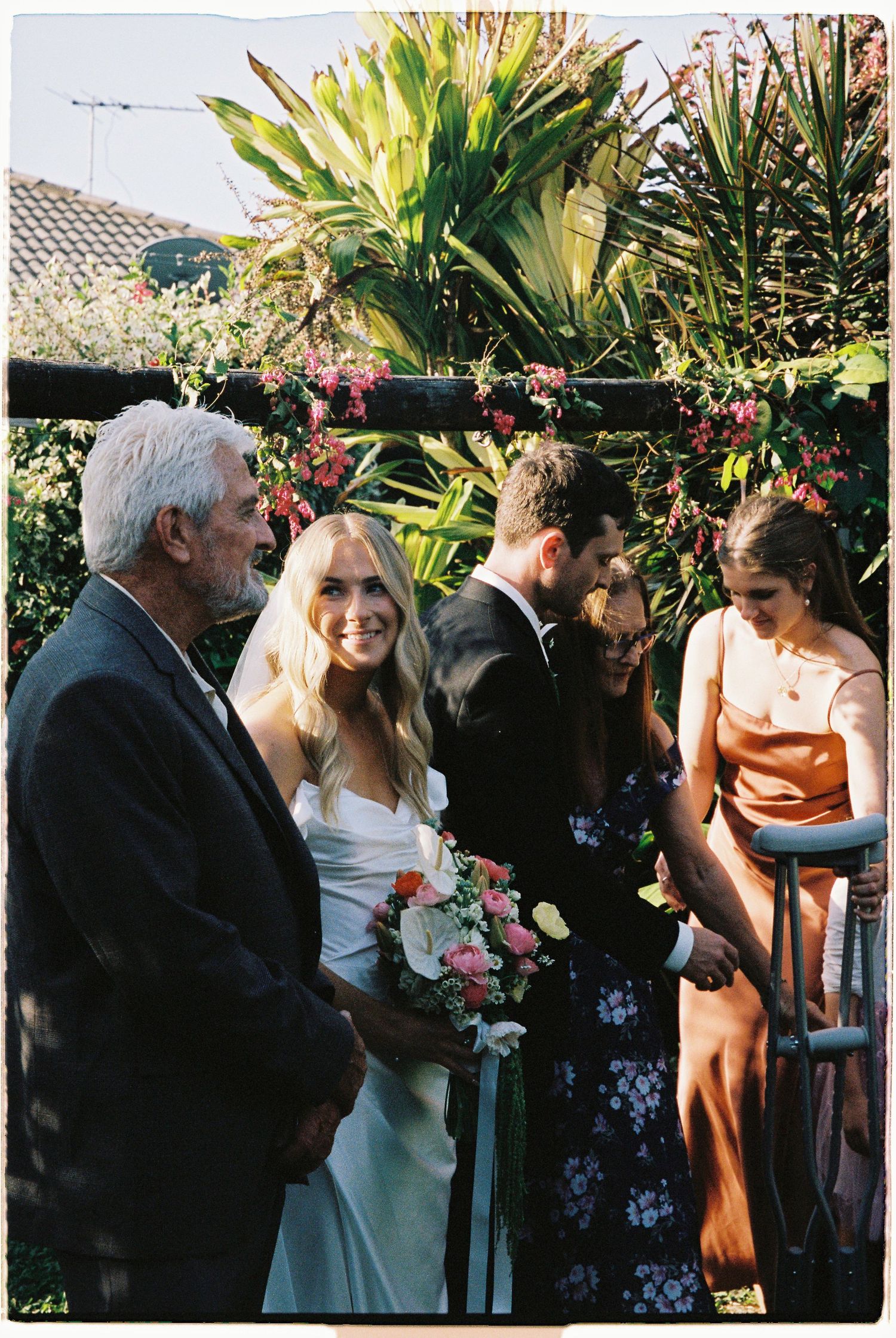 Smiling bride holding bouquet stands at outdoor ceremony altar surrounded by tropical plants and wedding guests.