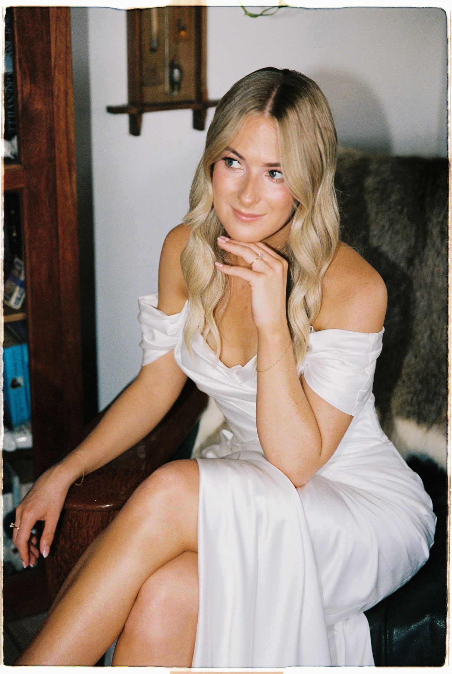 Blonde bride in white off-shoulder wedding dress seated, posing with hand on chin.