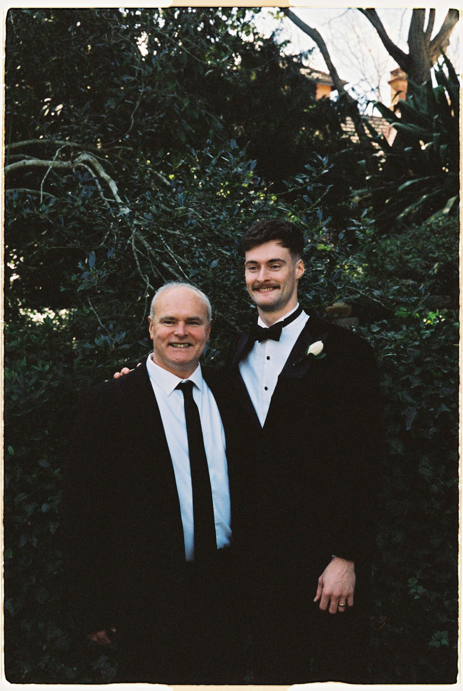 A groom in a black tuxedo poses with an older man in a garden setting.