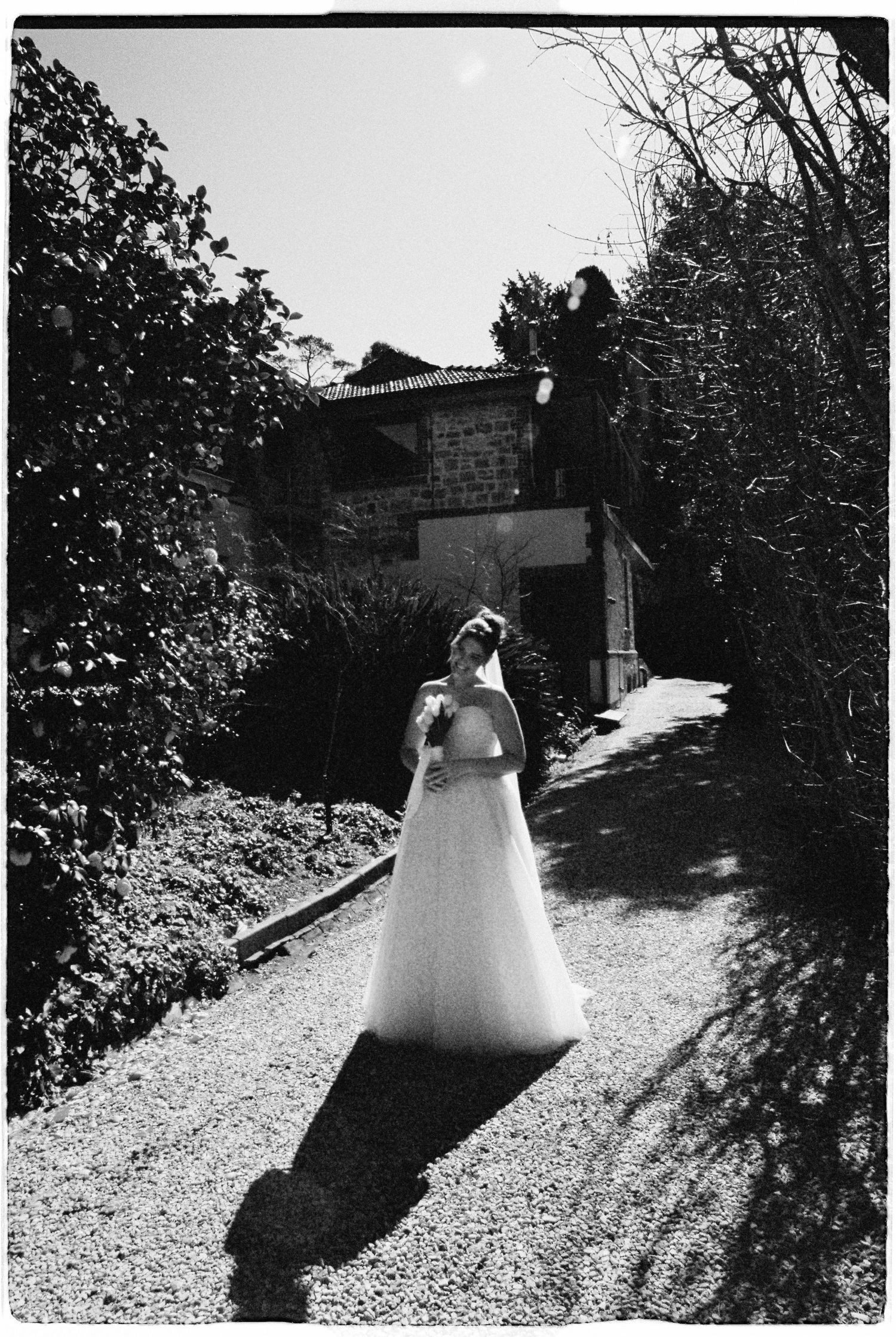 Bride in white gown walks garden path at rustic venue while photographer captures the moment from above.