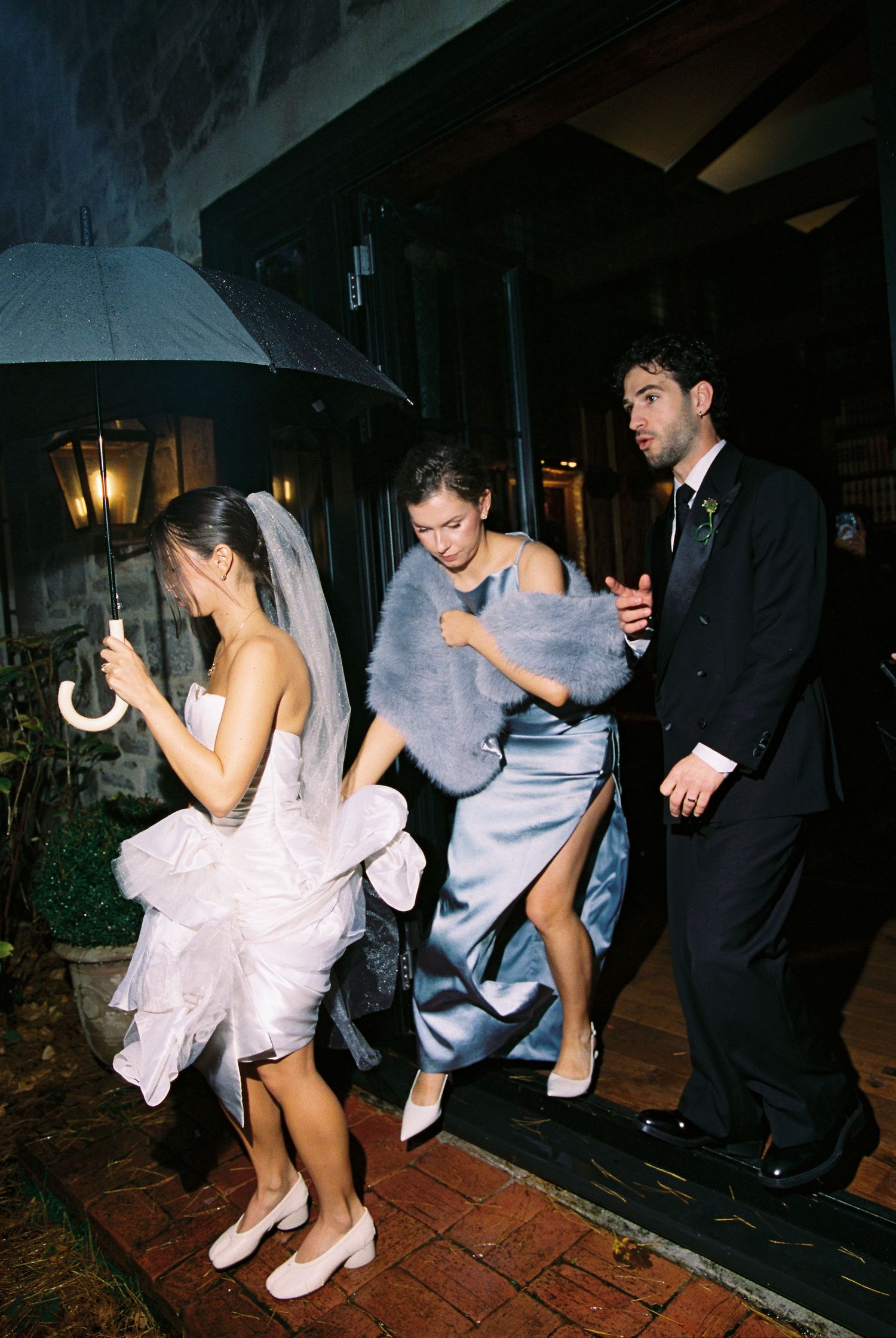 Bride with umbrella and bridesmaid in blue gown rushing through rain with groomsman.