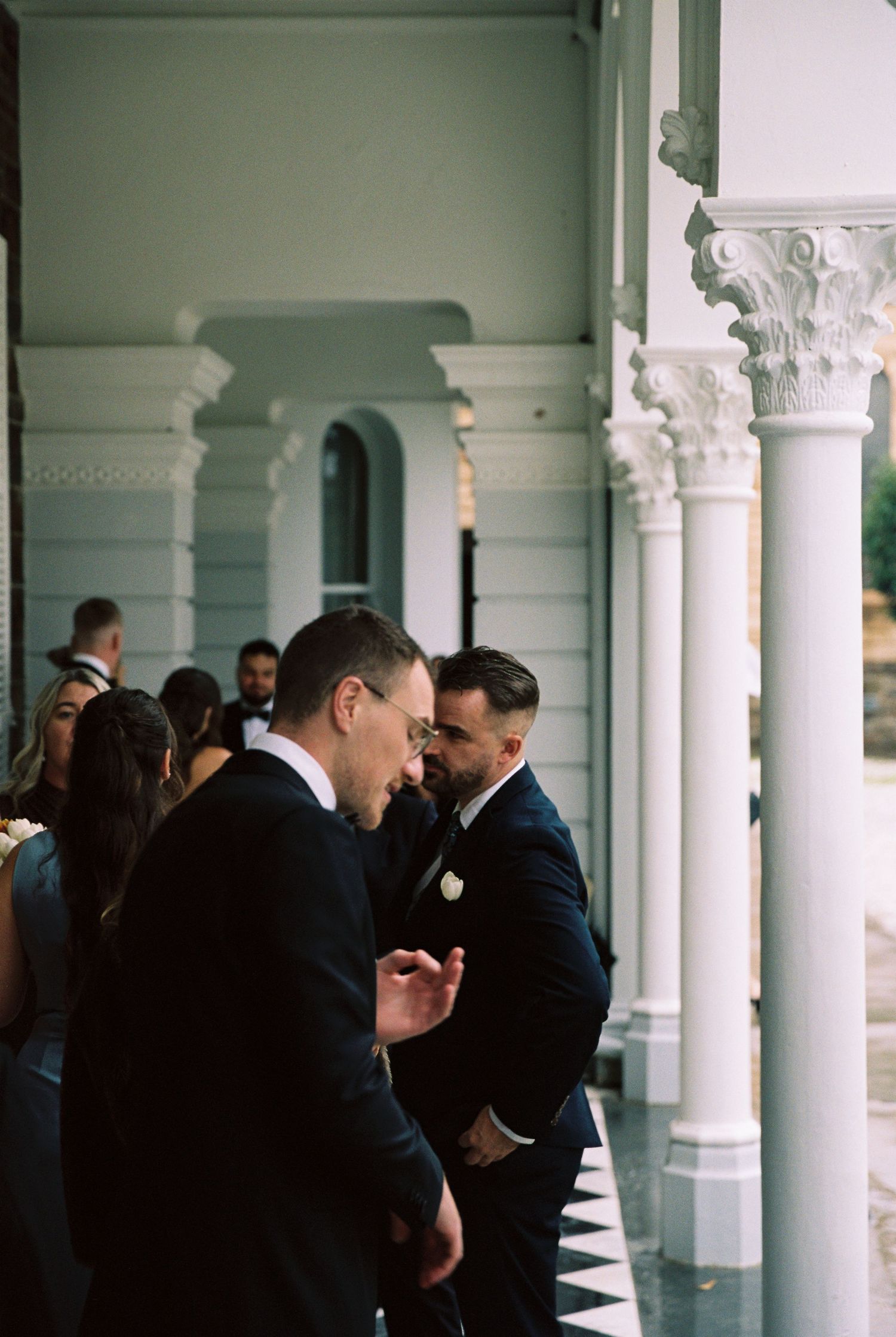 Two grooms in black suits share a moment on a white columned wedding venue veranda.