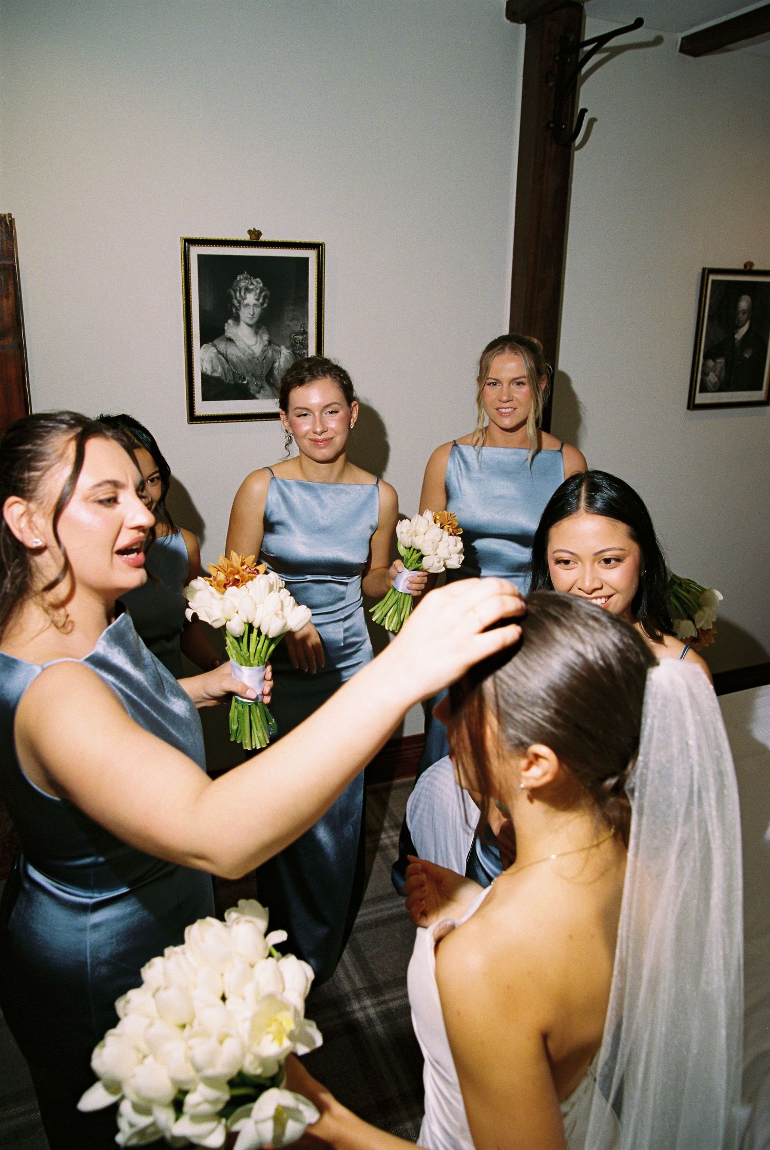 Bridesmaids in blue satin dresses help bride with veil while holding white floral bouquets.