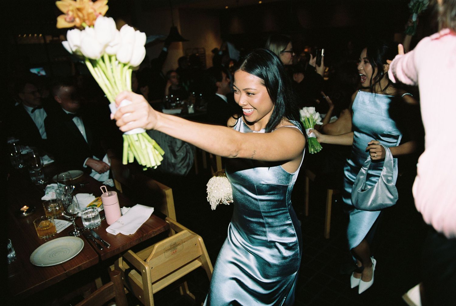Bridesmaid in silver satin dress holds bouquet of white and peach flowers at wedding reception.