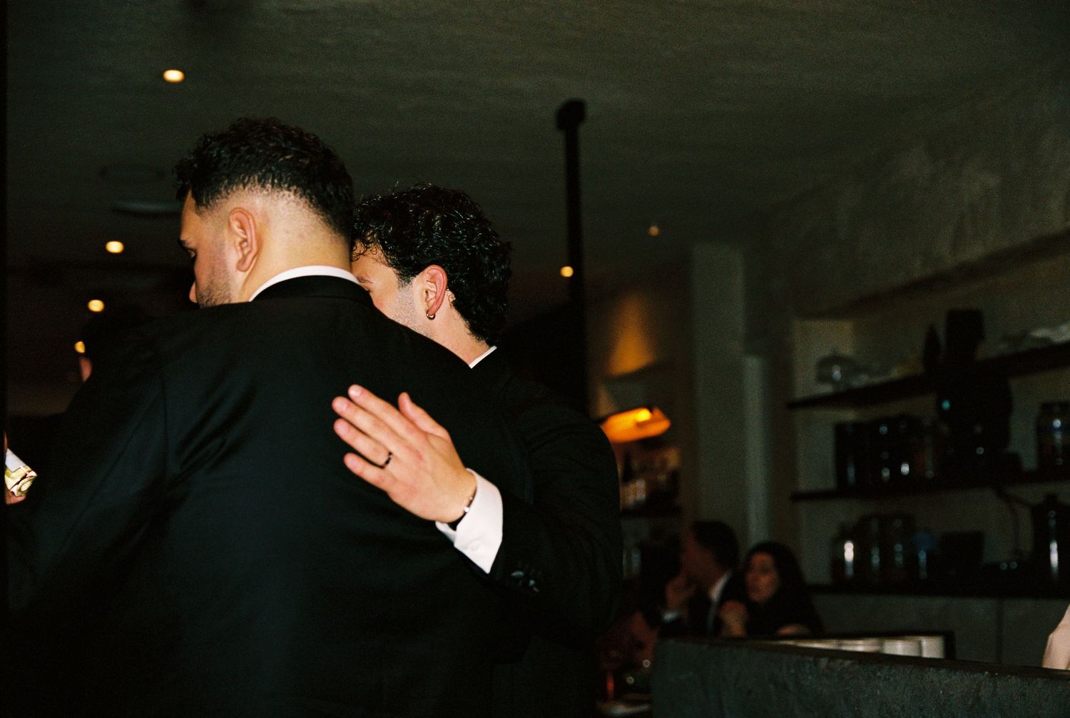 Two men in formal black suits embracing at a wedding reception bar.