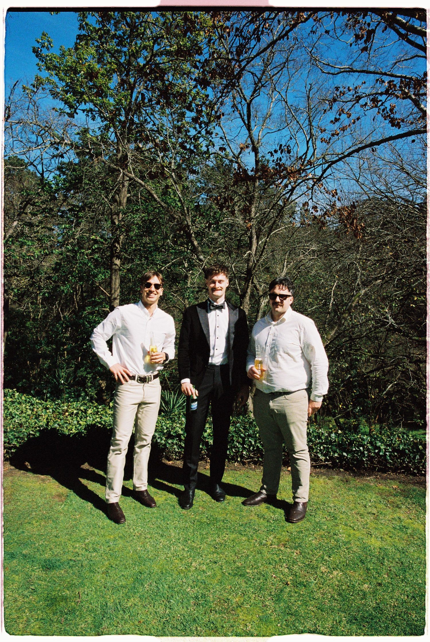 Three men in formal attire holding drinks pose outdoors in a sunny garden with lush green trees.