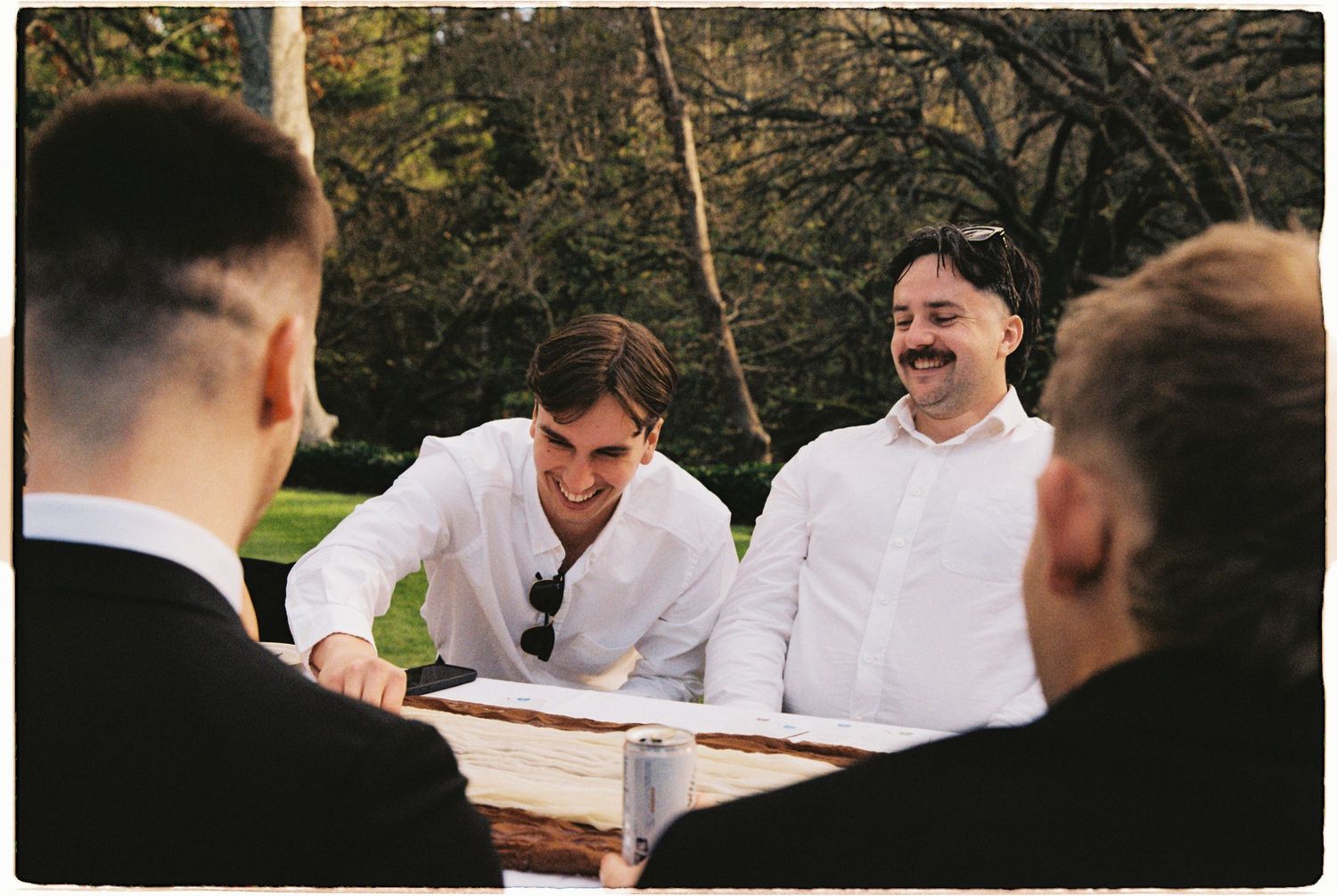 Groomsmen laugh and gather around a wooden table outdoors during a relaxed wedding celebration.