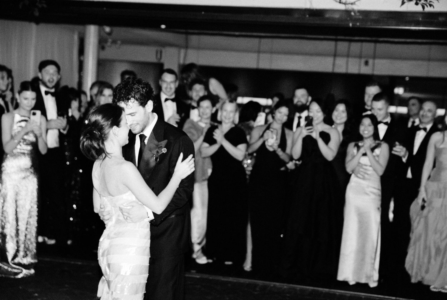 Black and white photo of couple sharing first dance at wedding reception with guests applauding in background.