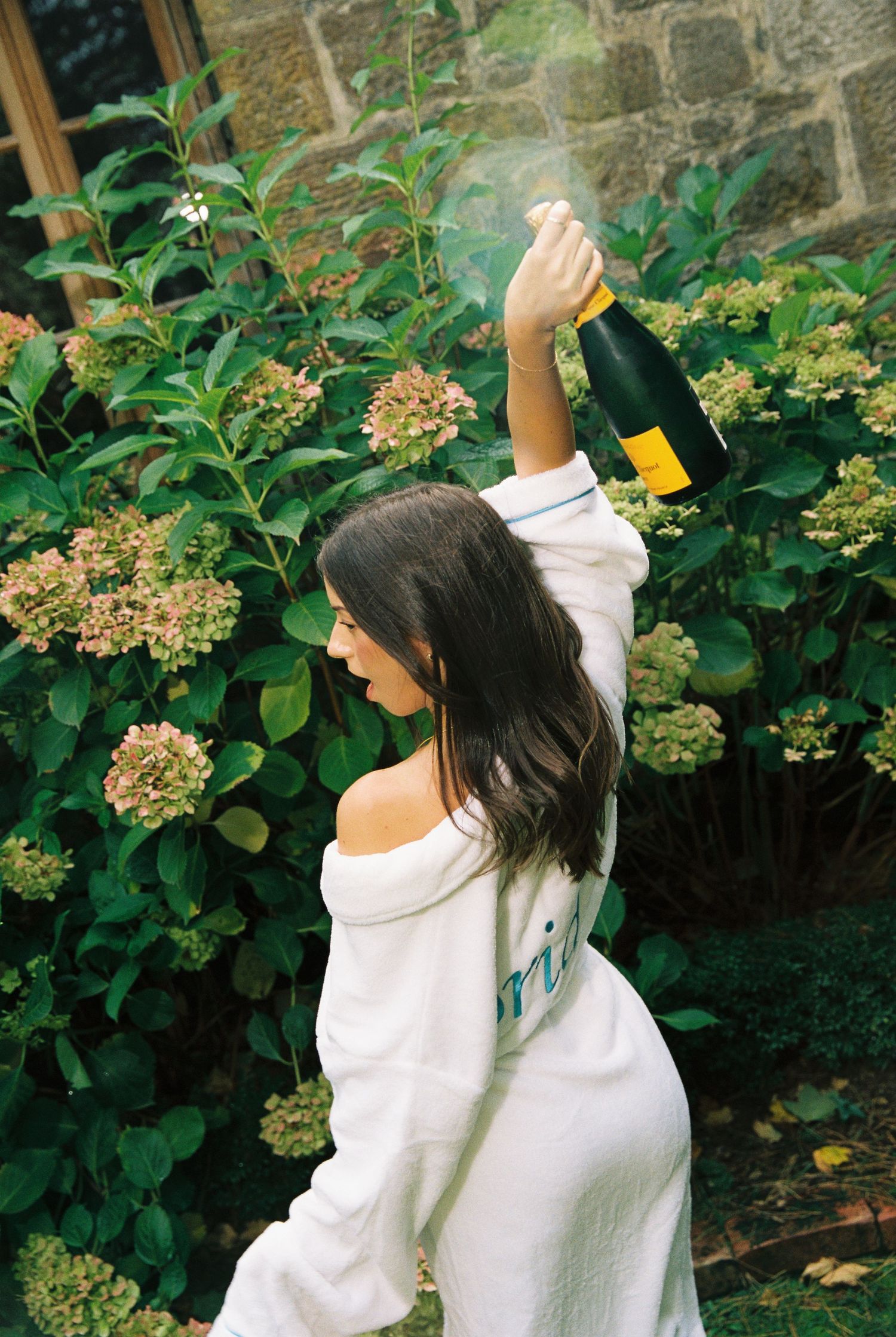 Woman in white robe popping champagne bottle outdoors near blooming hydrangea bushes.
