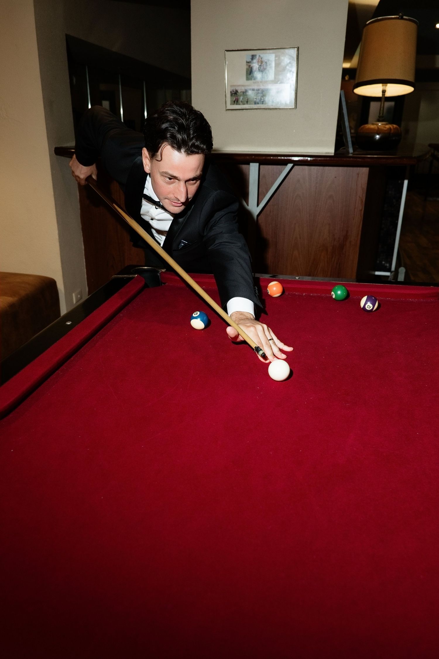 Elegant man in tuxedo leaning over red pool table preparing to take a shot at billiard balls.
