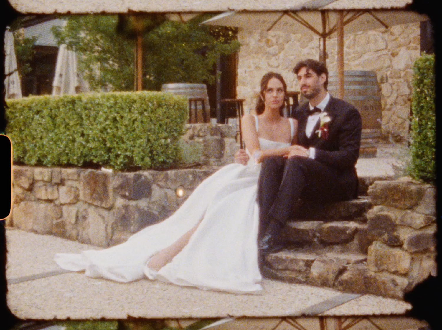Bride and groom in wedding attire sitting on stone steps at rustic outdoor venue.