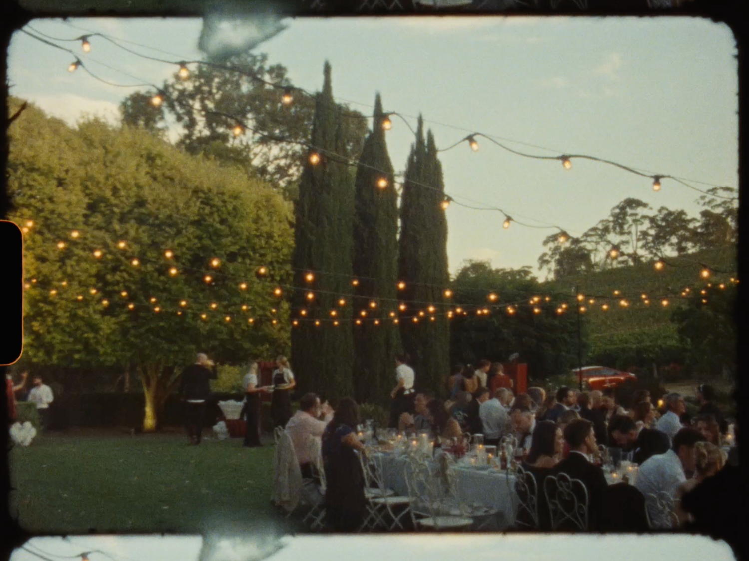 Elegant outdoor wedding reception at dusk with guests dining under warm string lights near cypress trees.