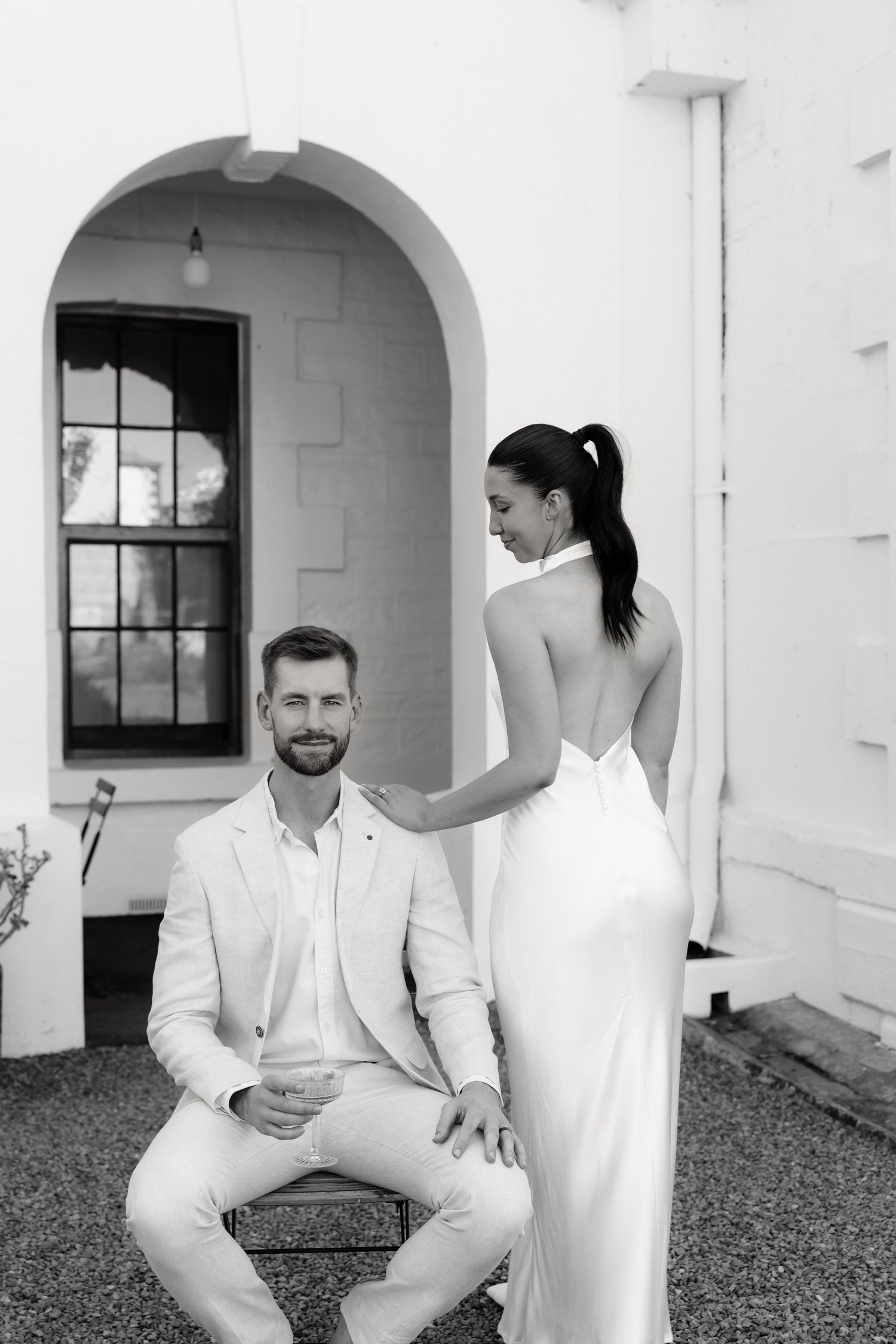 Black and white portrait of groom seated as bride in backless gown stands beside him.