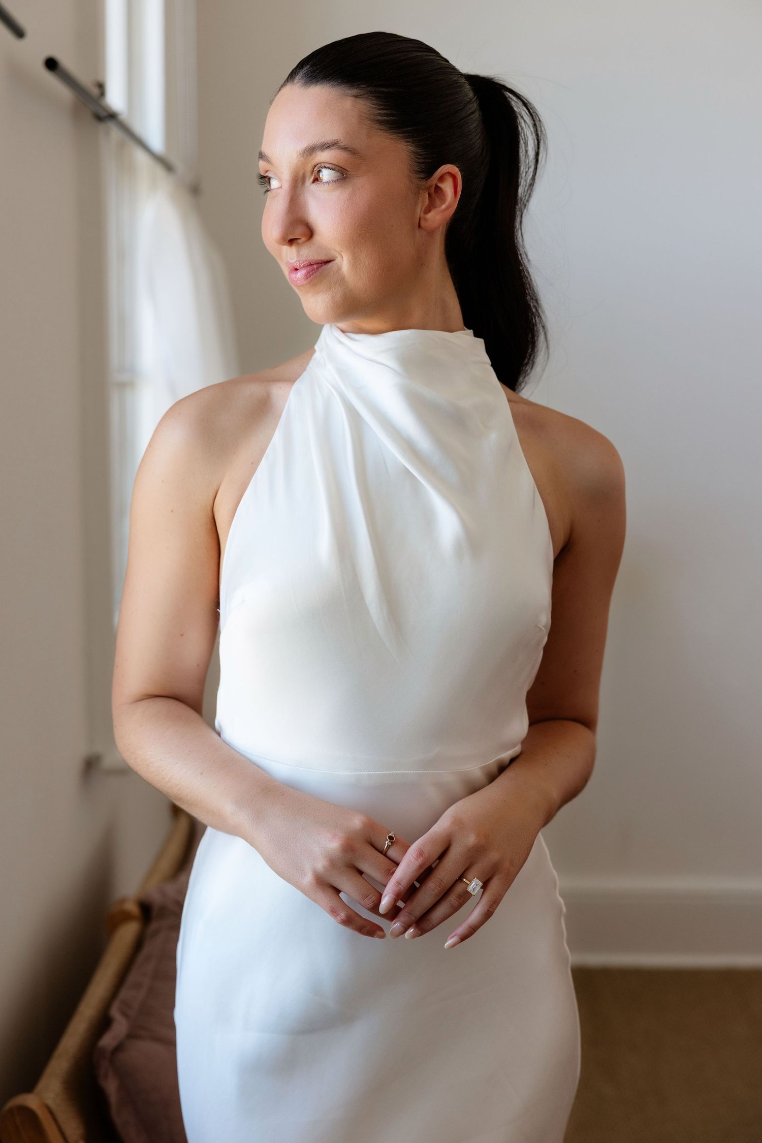 Dark-haired bride in white halter neck wedding dress gazing sideways in a softly lit room.