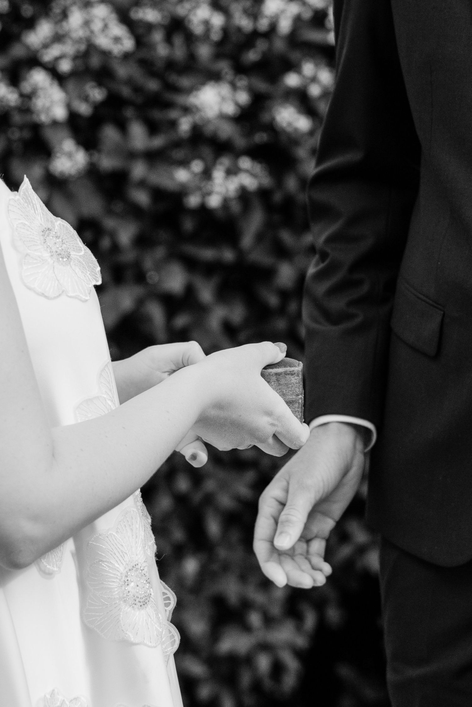 Black and white photo of bride placing wedding ring on groom's finger during outdoor ceremony.