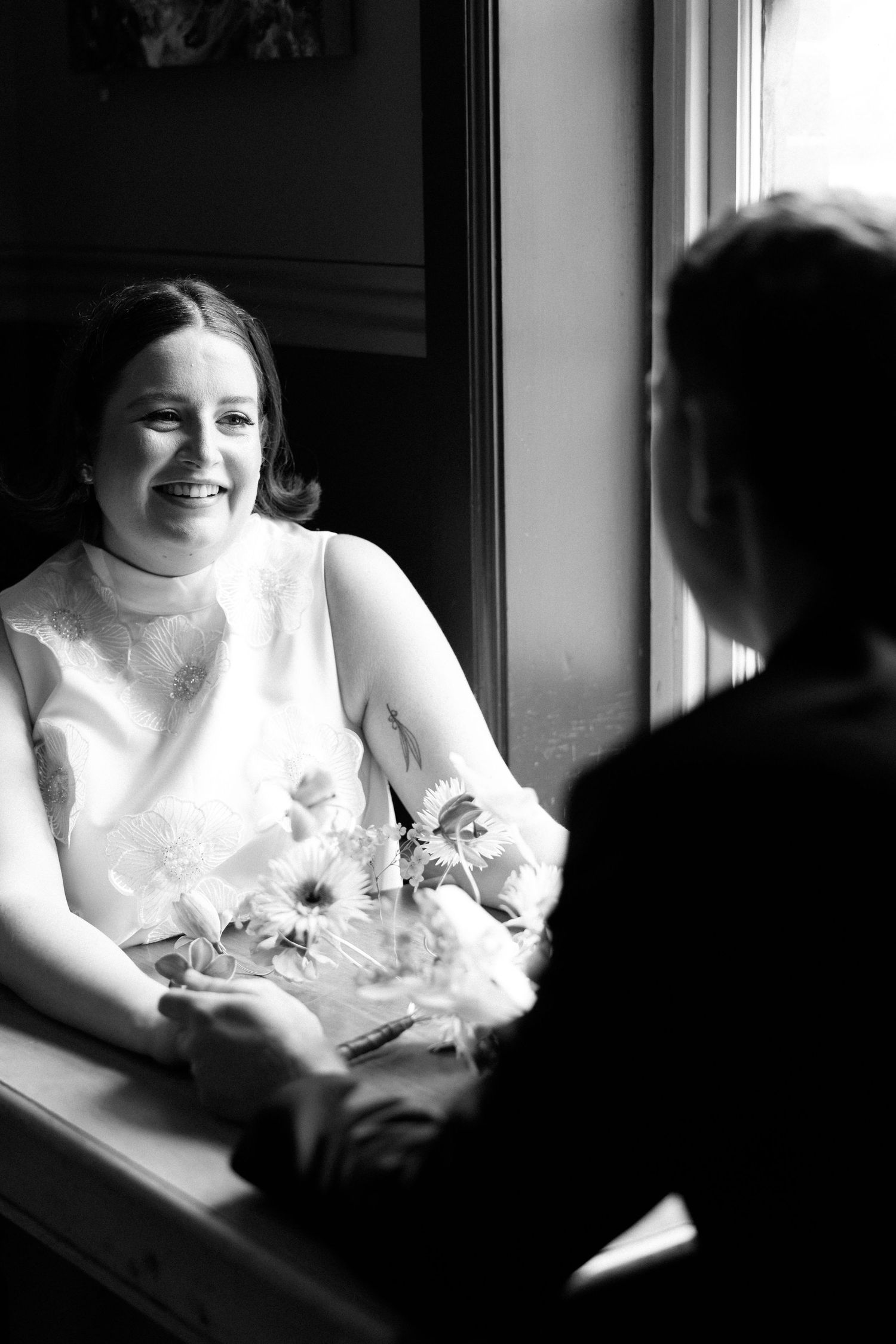 Black and white photo of a smiling bride holding flowers, sitting by a window, holding hands with groom.