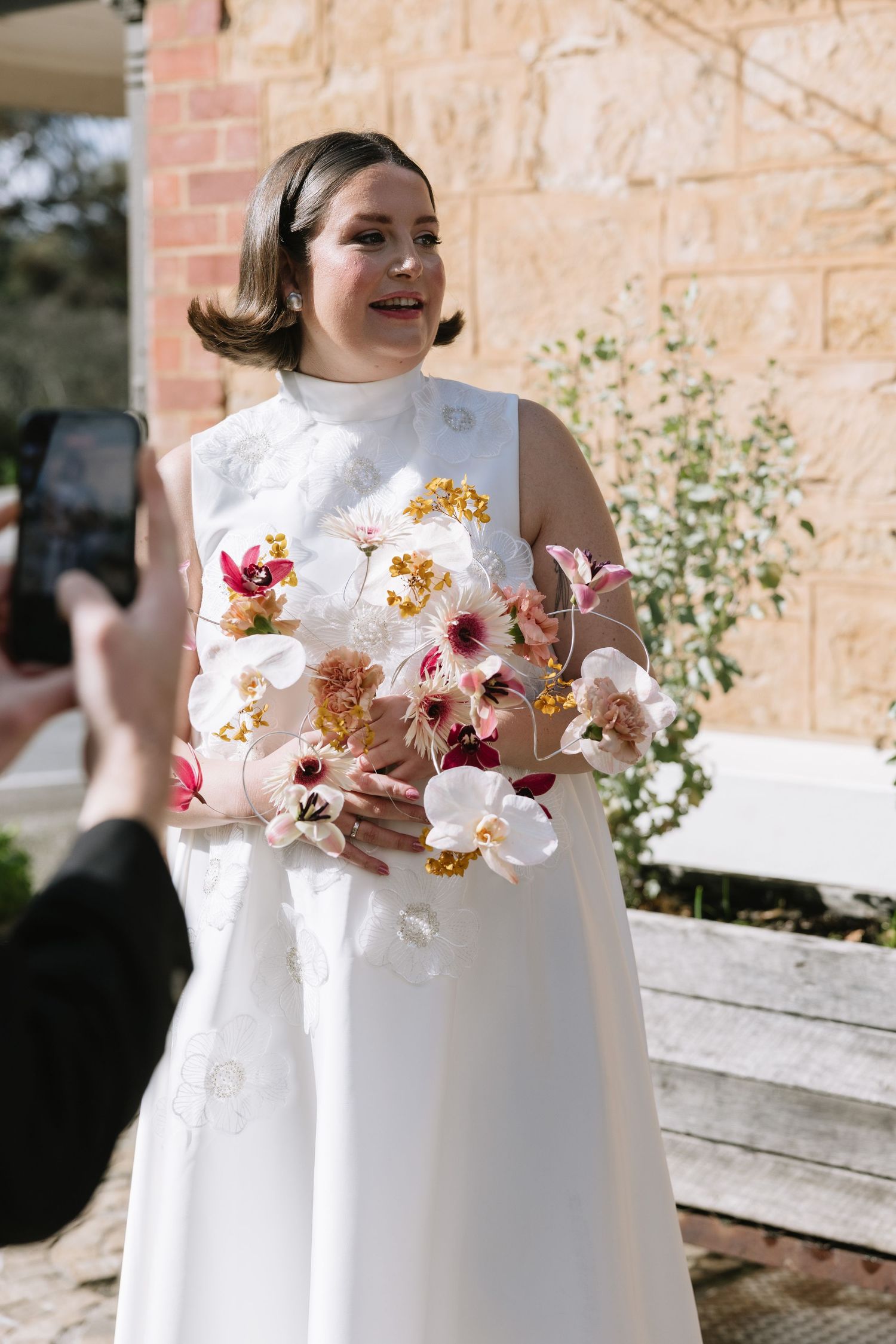 Bride in white sleeveless gown holding colorful orchid bouquet while being photographed outdoors near stone building.