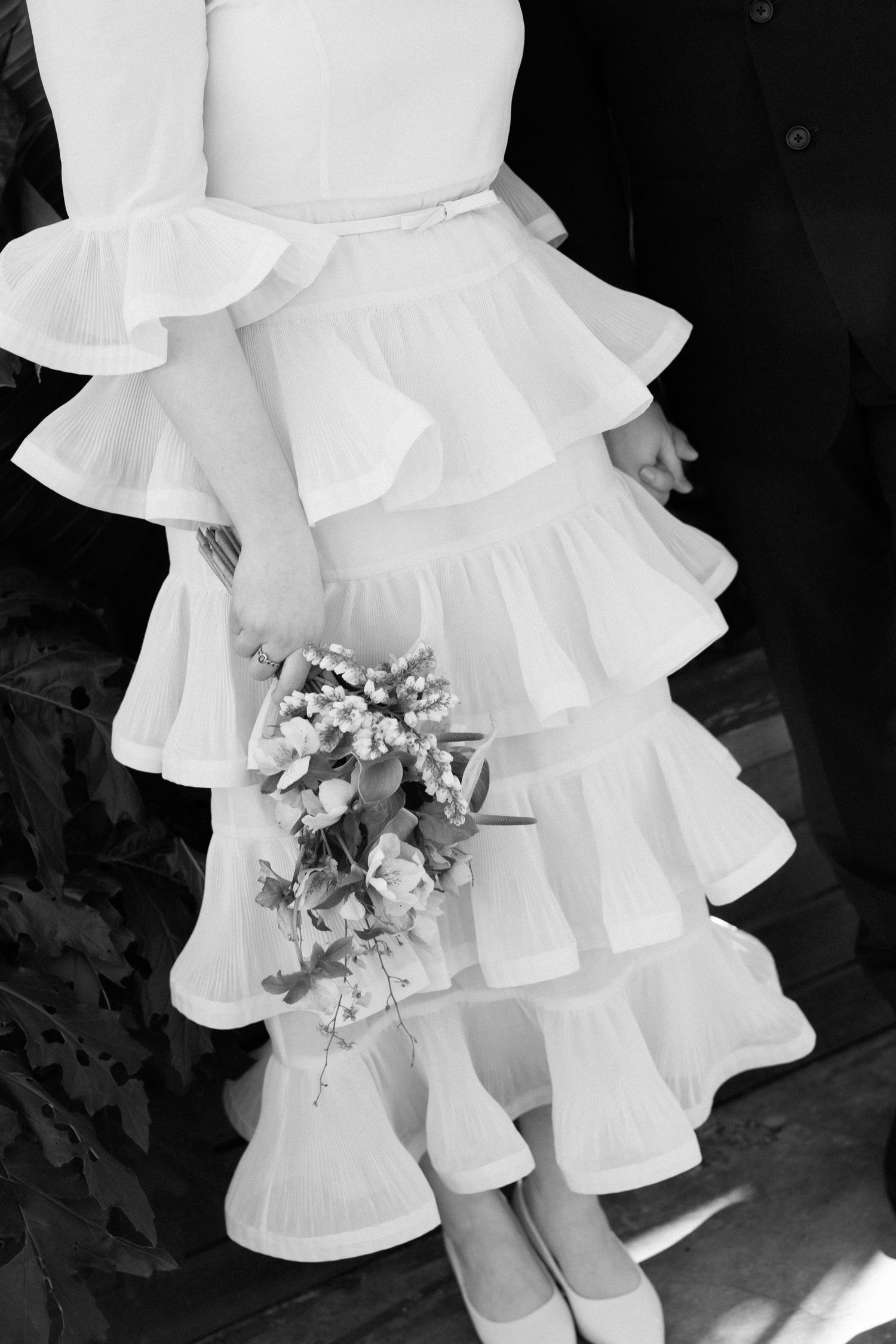 Bride in tiered ruffle white wedding dress holding a small floral bouquet, black and white photo.