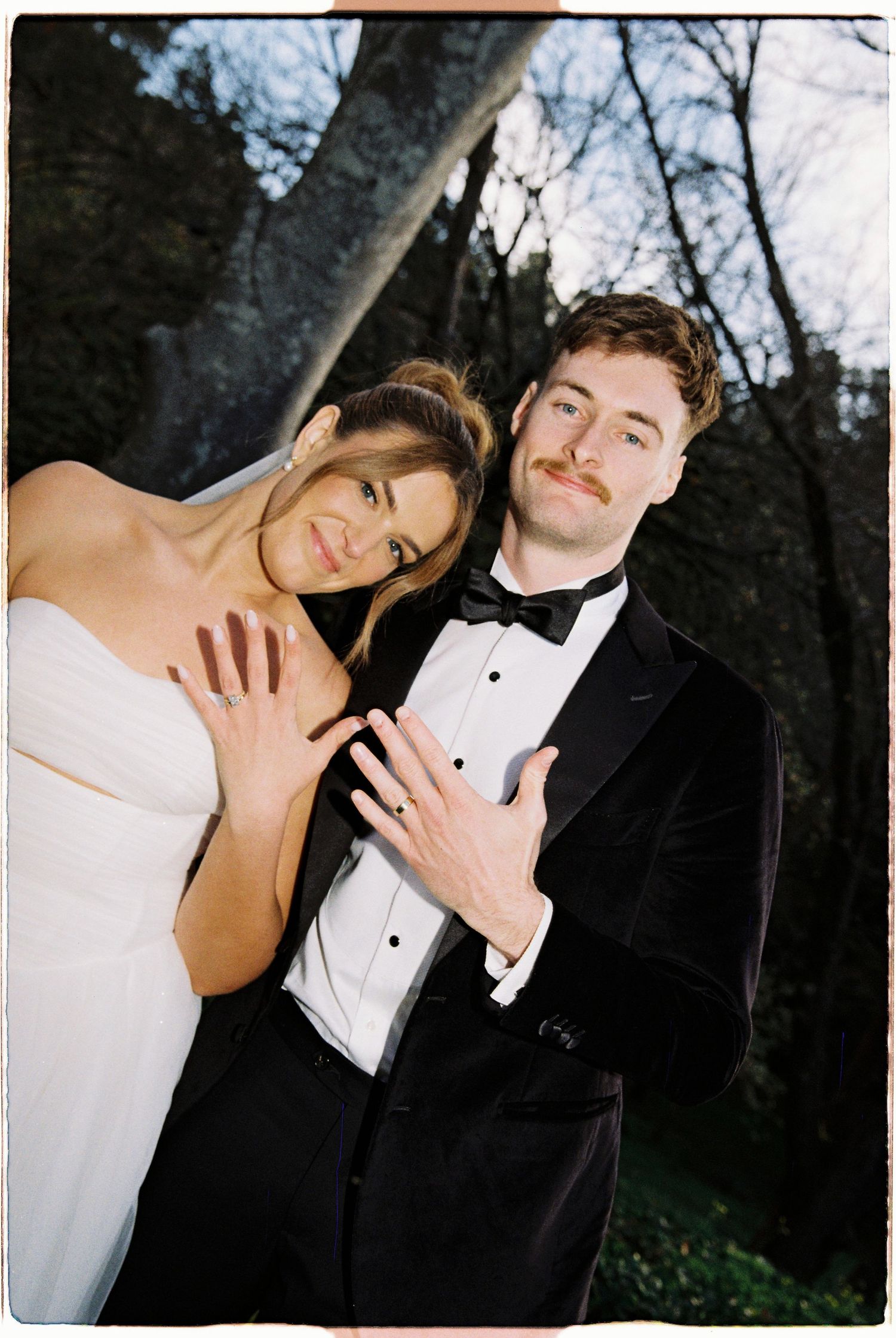 Bride and groom showing off wedding rings outdoors, wearing white gown and black tuxedo.