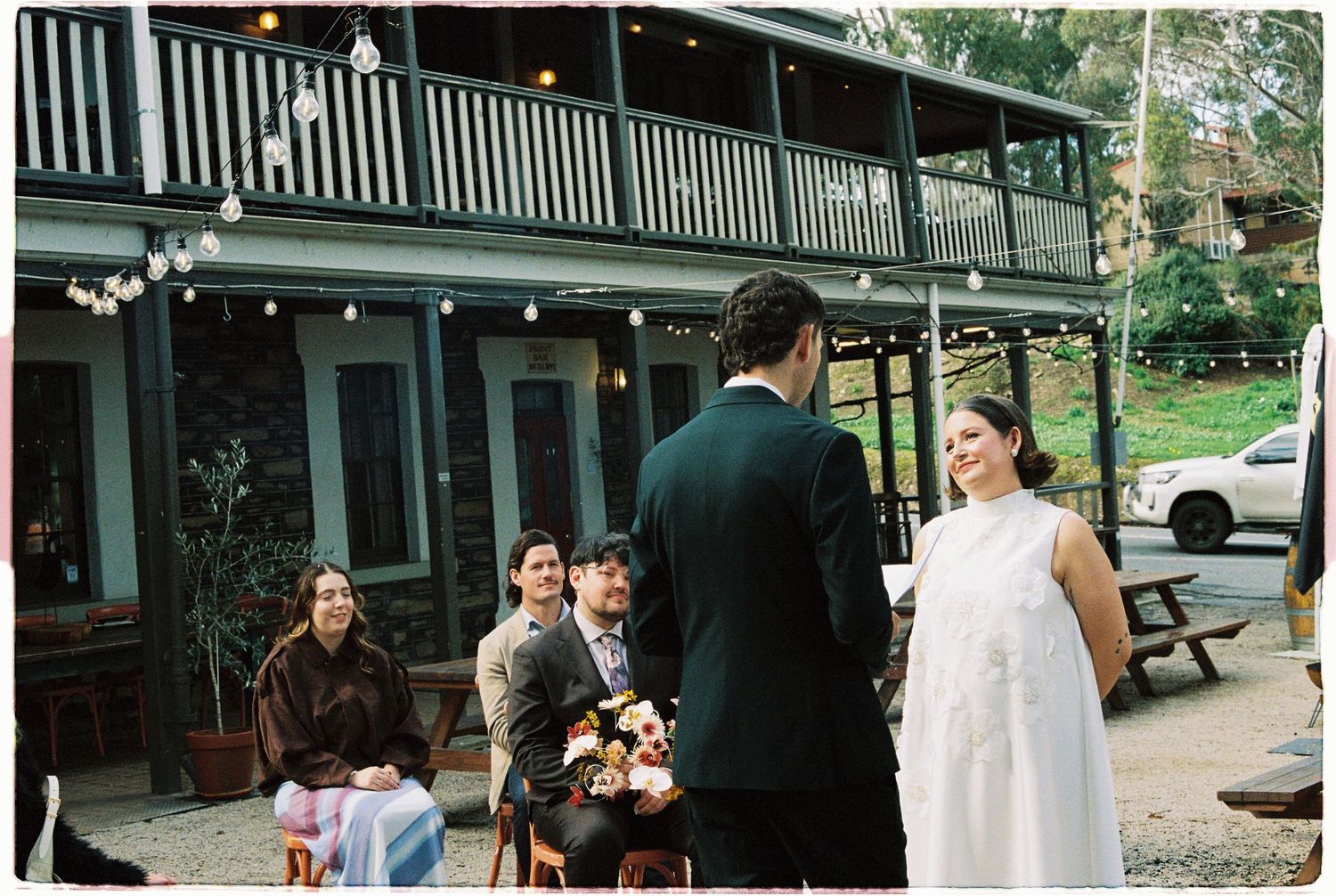 Outdoor wedding ceremony at rustic venue with string lights, groom in suit facing bride in white dress with guests seated.
