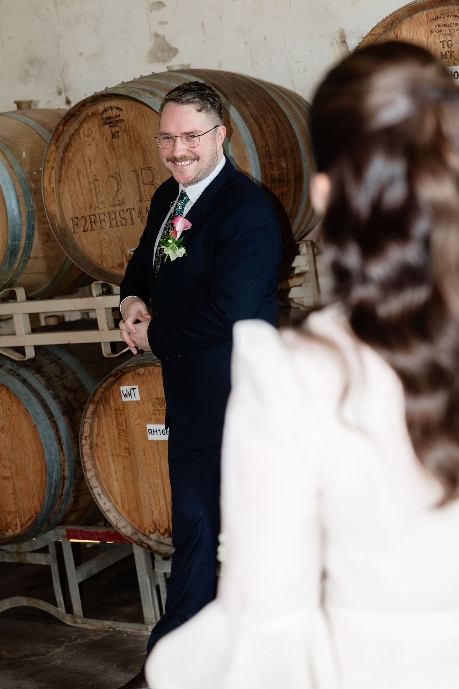 Groom in navy suit smiles at bride near rustic wine barrels in a winery wedding venue.