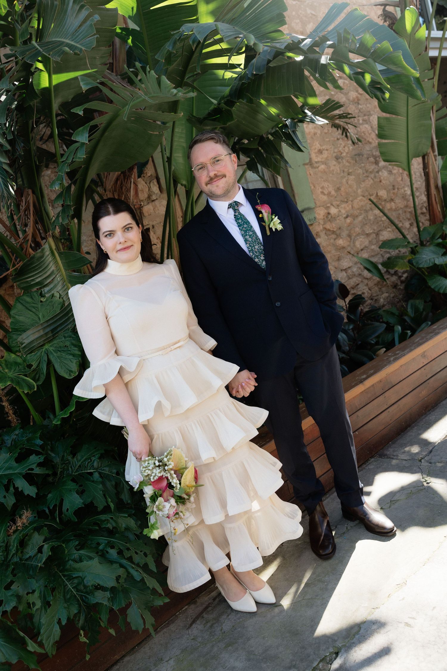 Bride in ruffled cream dress holds bouquet beside groom in navy suit with floral boutonniere, posing among tropical greenery.