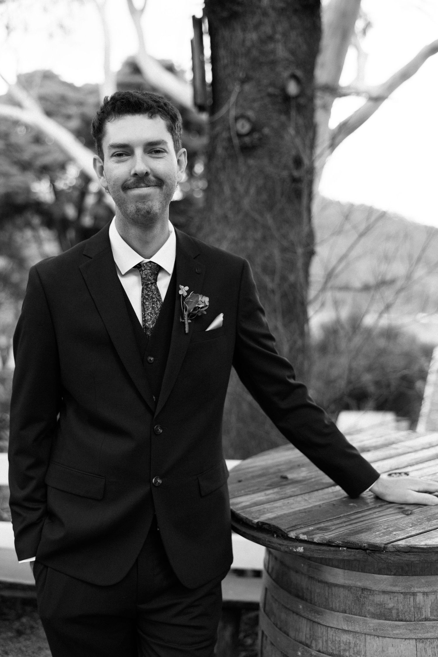 Black and white portrait of a smiling groom in a suit with boutonniere, leaning on a wooden barrel outdoors.