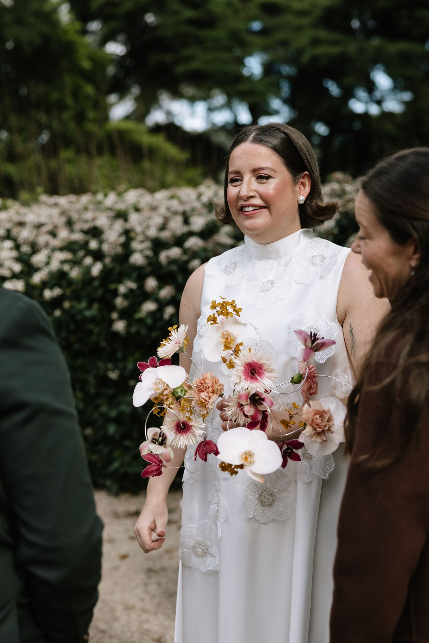 Smiling bride in white gown holding colorful floral bouquet with orchids outdoors near lush greenery.