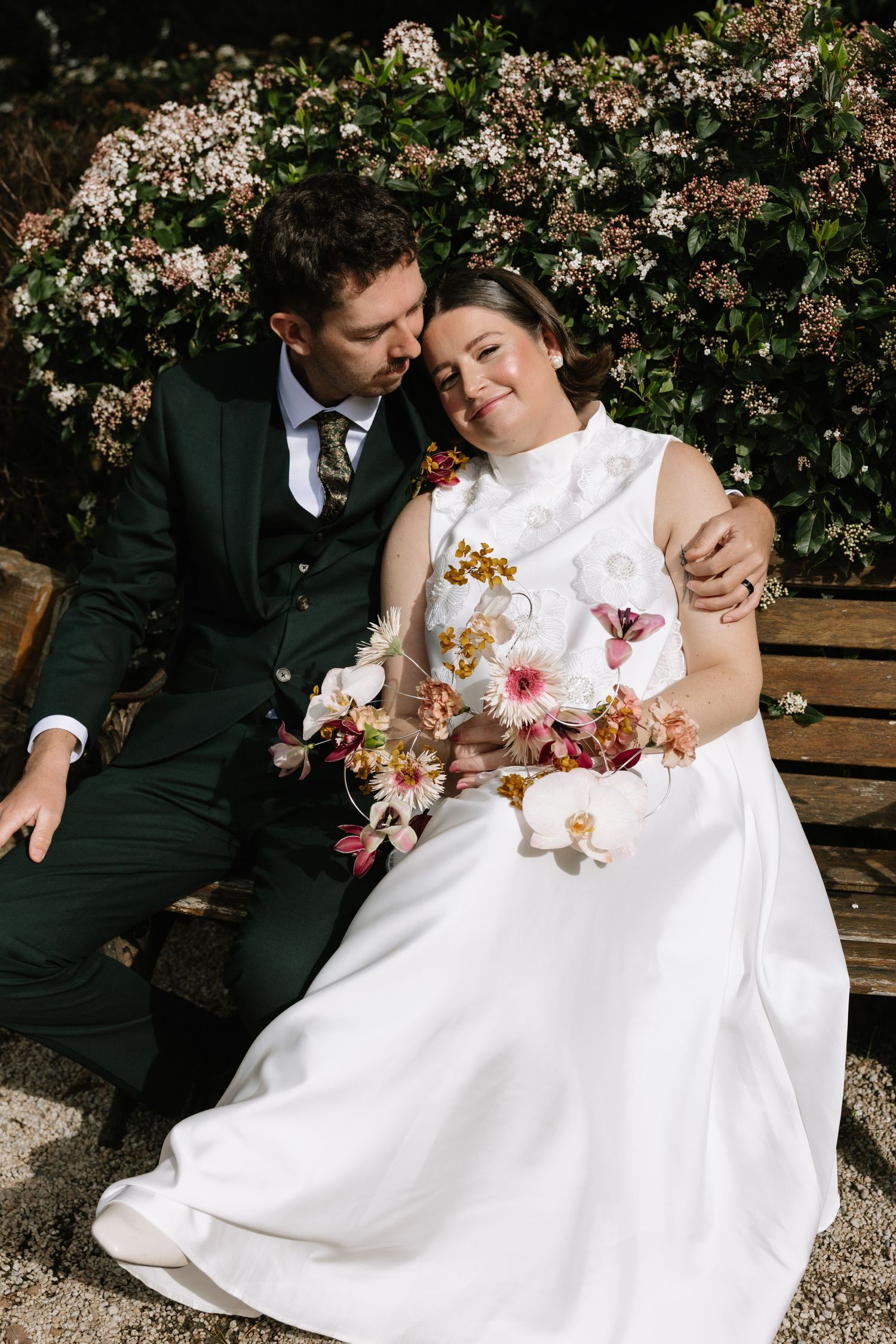 Bride and groom sitting on a bench surrounded by blooming flowers, bride holding a colorful bouquet in wedding attire.