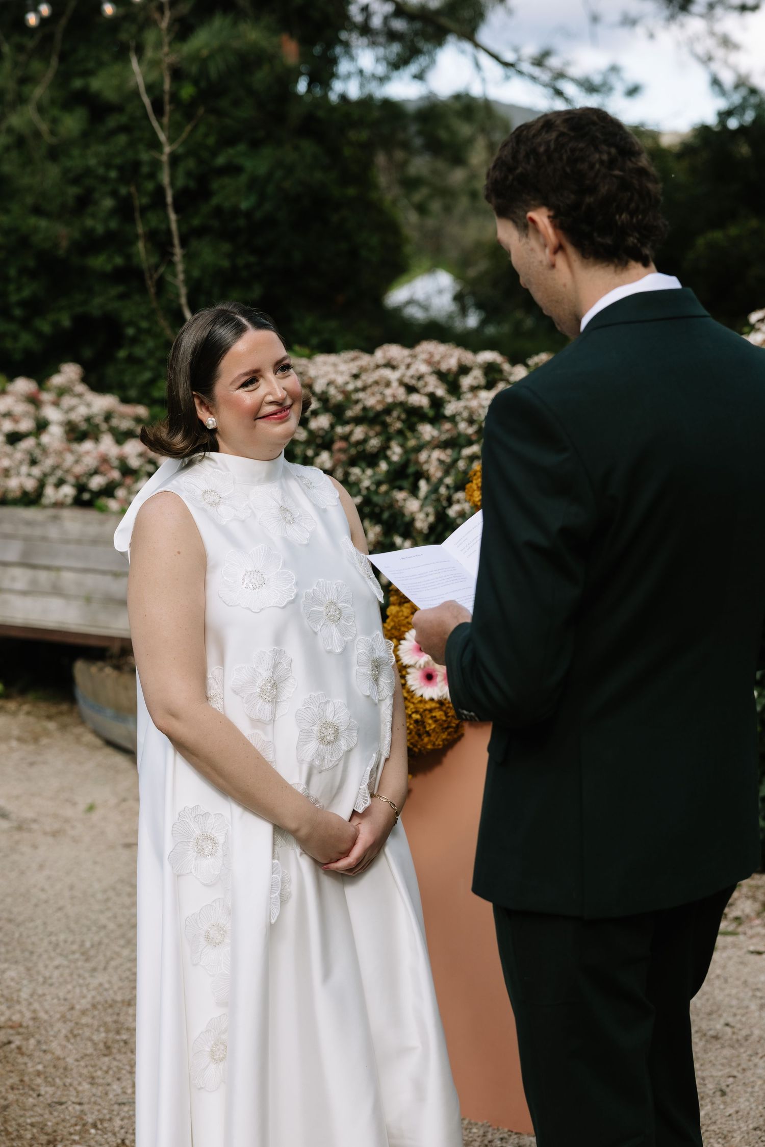 Bride in white embroidered gown smiles at groom in dark suit during outdoor wedding ceremony with floral backdrop.