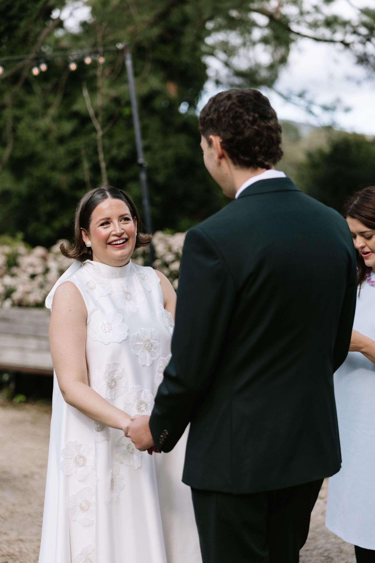 Bride in white lace dress smiling joyfully at groom during outdoor wedding ceremony while holding hands.