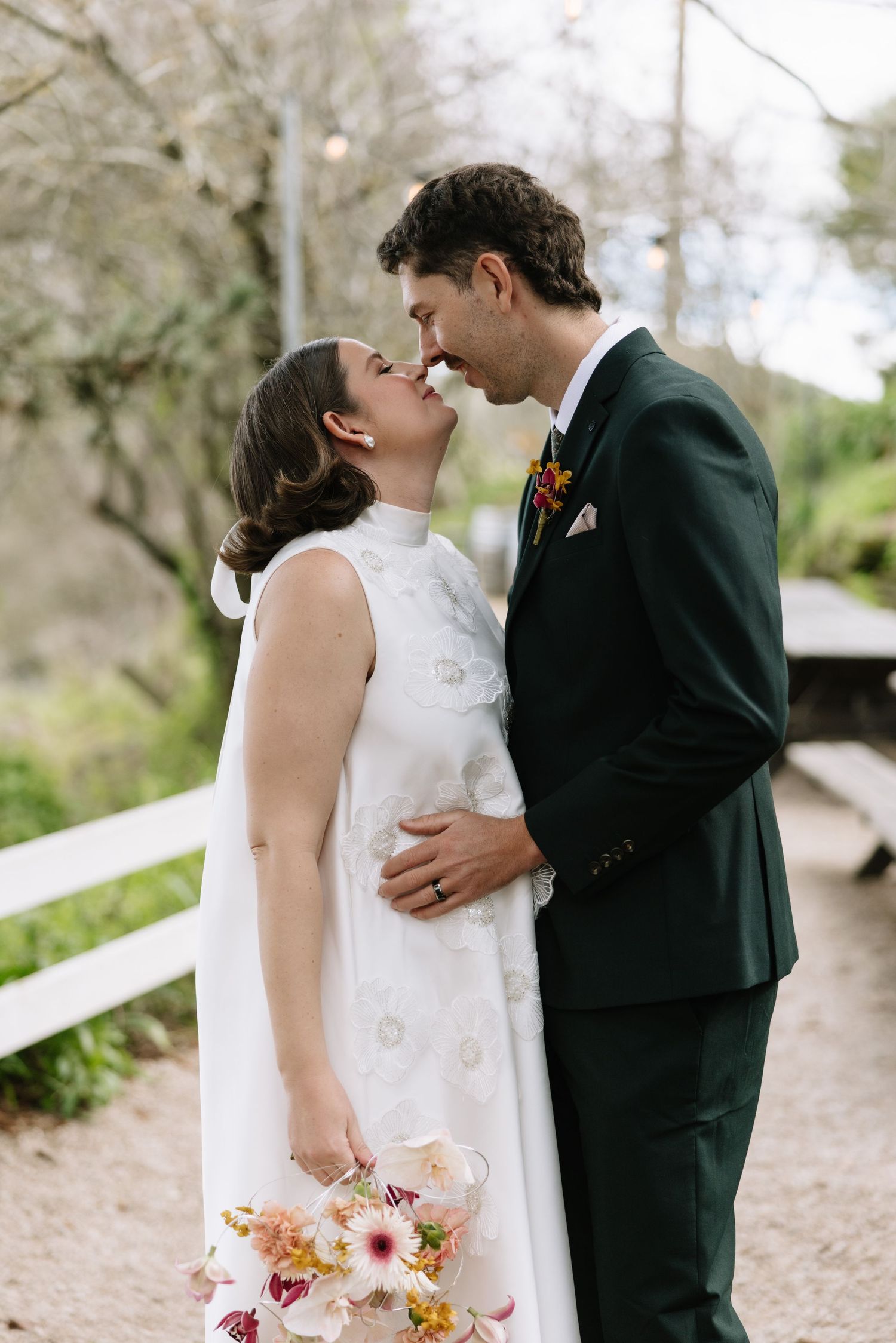 A bride and groom share a tender moment, nearly kissing outdoors, she in a white gown holding a bouquet, he in a dark green suit.