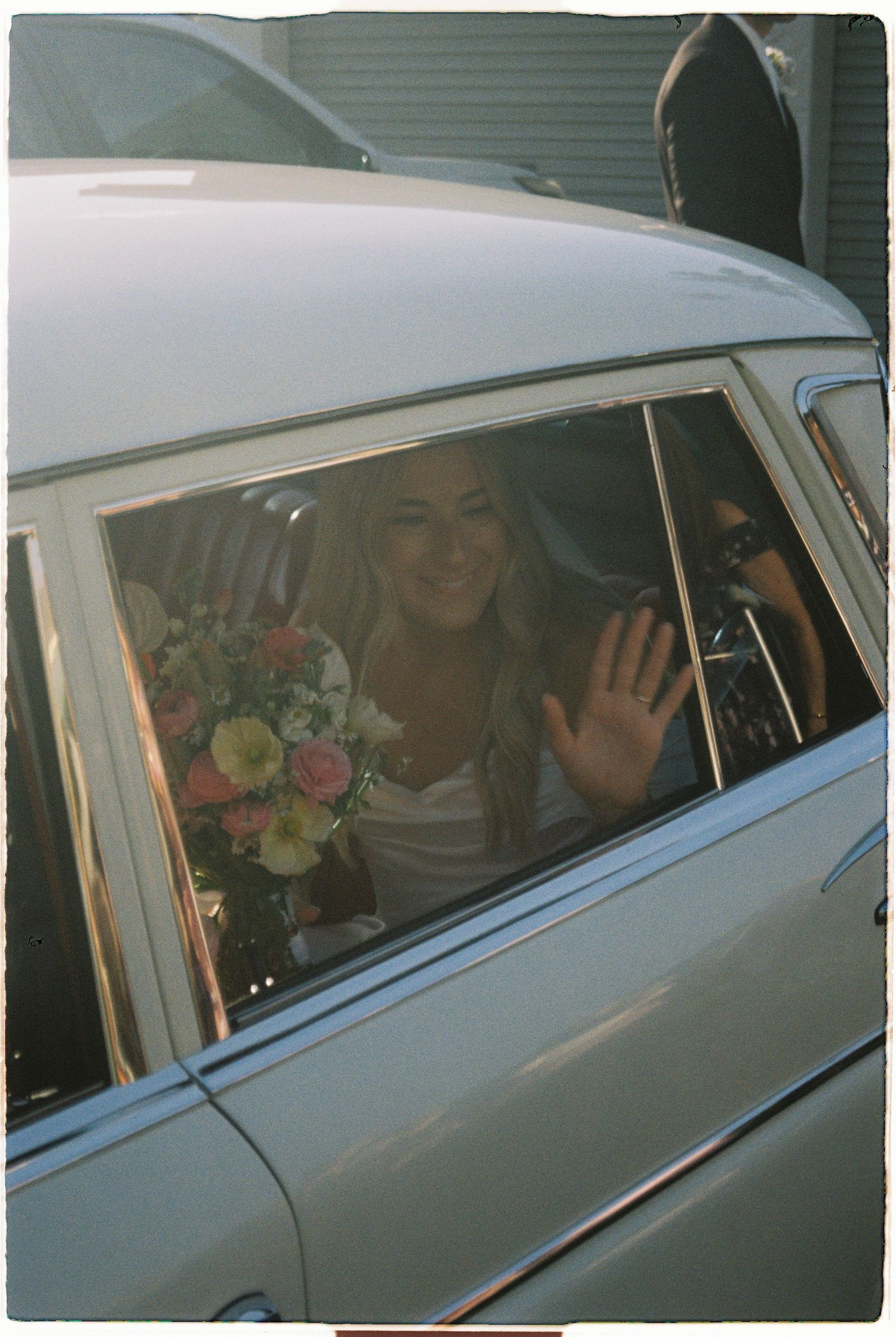 Smiling bride in vintage car waving with colorful bridal bouquet on wedding day.