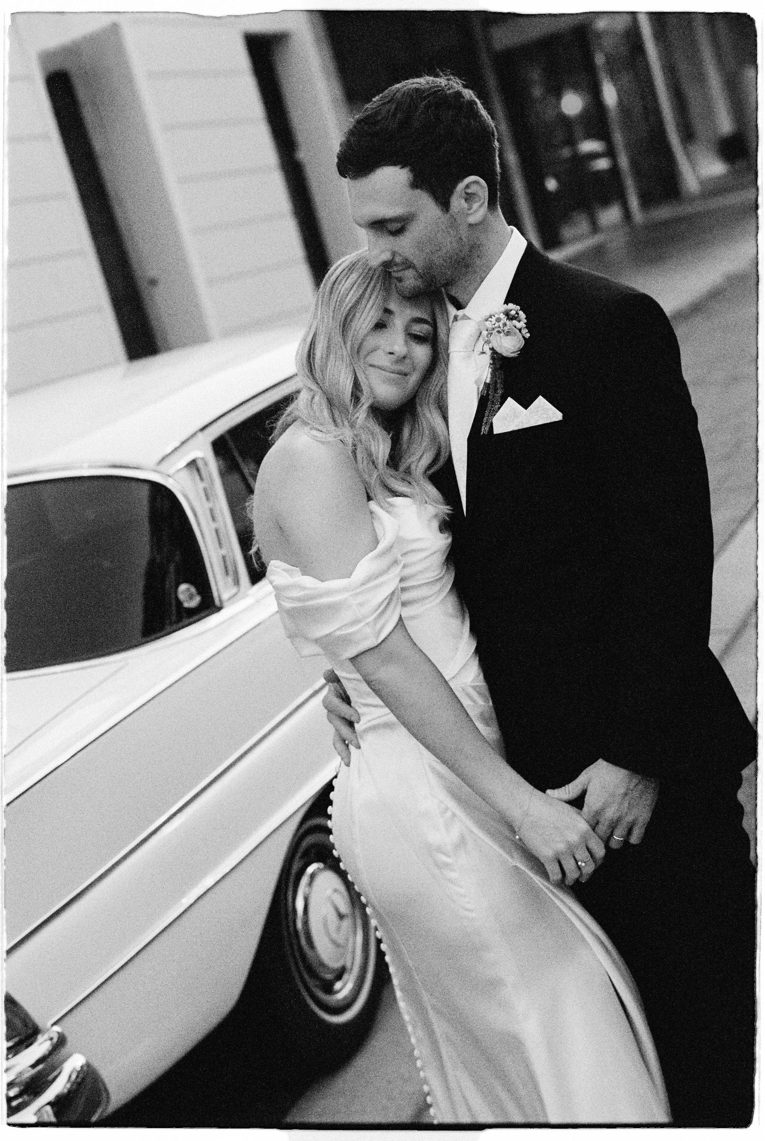 Bride and groom embracing tenderly beside a vintage white car in elegant black and white photo.