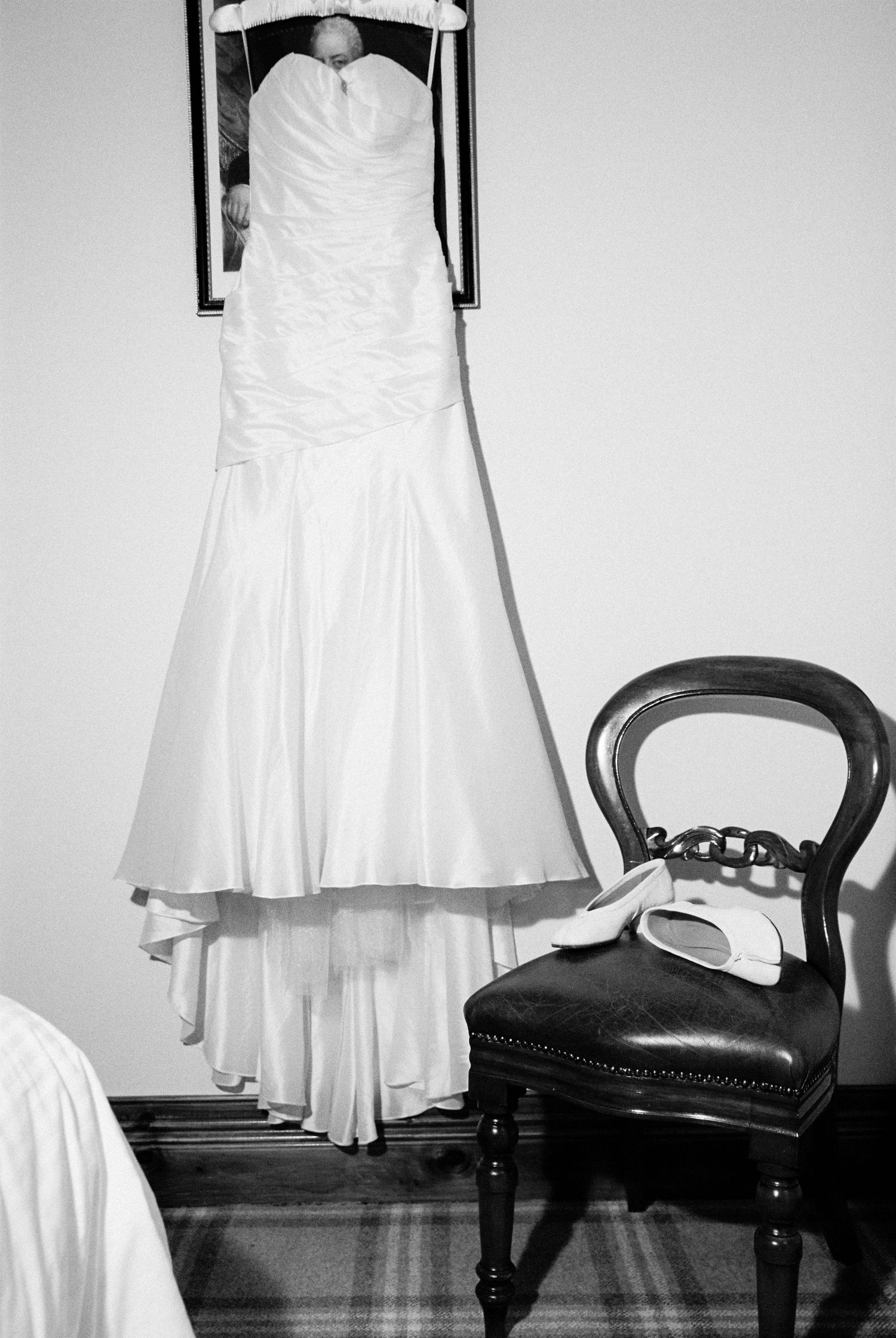 Elegant strapless mermaid wedding dress hanging in room beside vintage chair with bridal shoes.