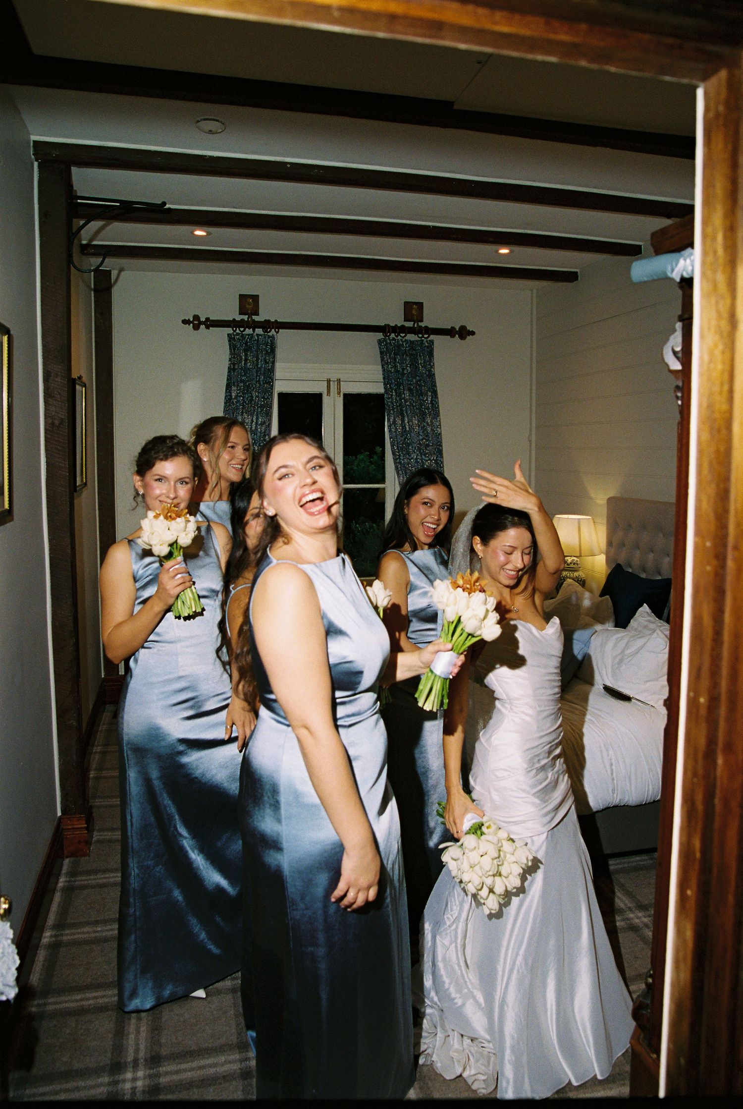 Bride and bridesmaids in blue satin dresses laughing while holding white bouquets.