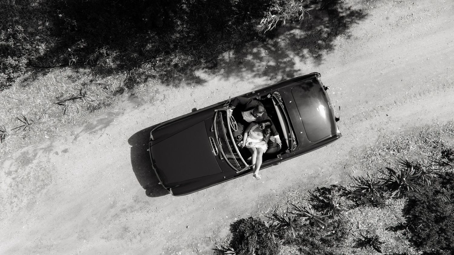 Aerial black and white view of bride sitting in convertible car on dirt road.