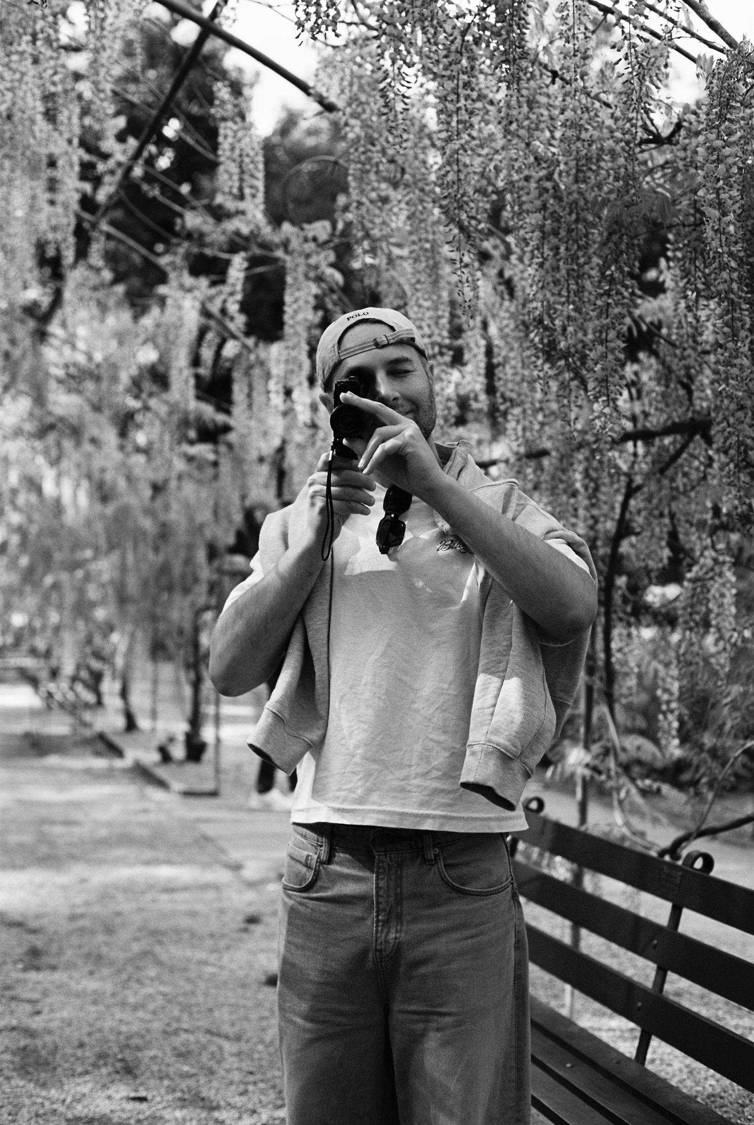 Young man photographing with camera amid hanging wisteria branches in black and white.