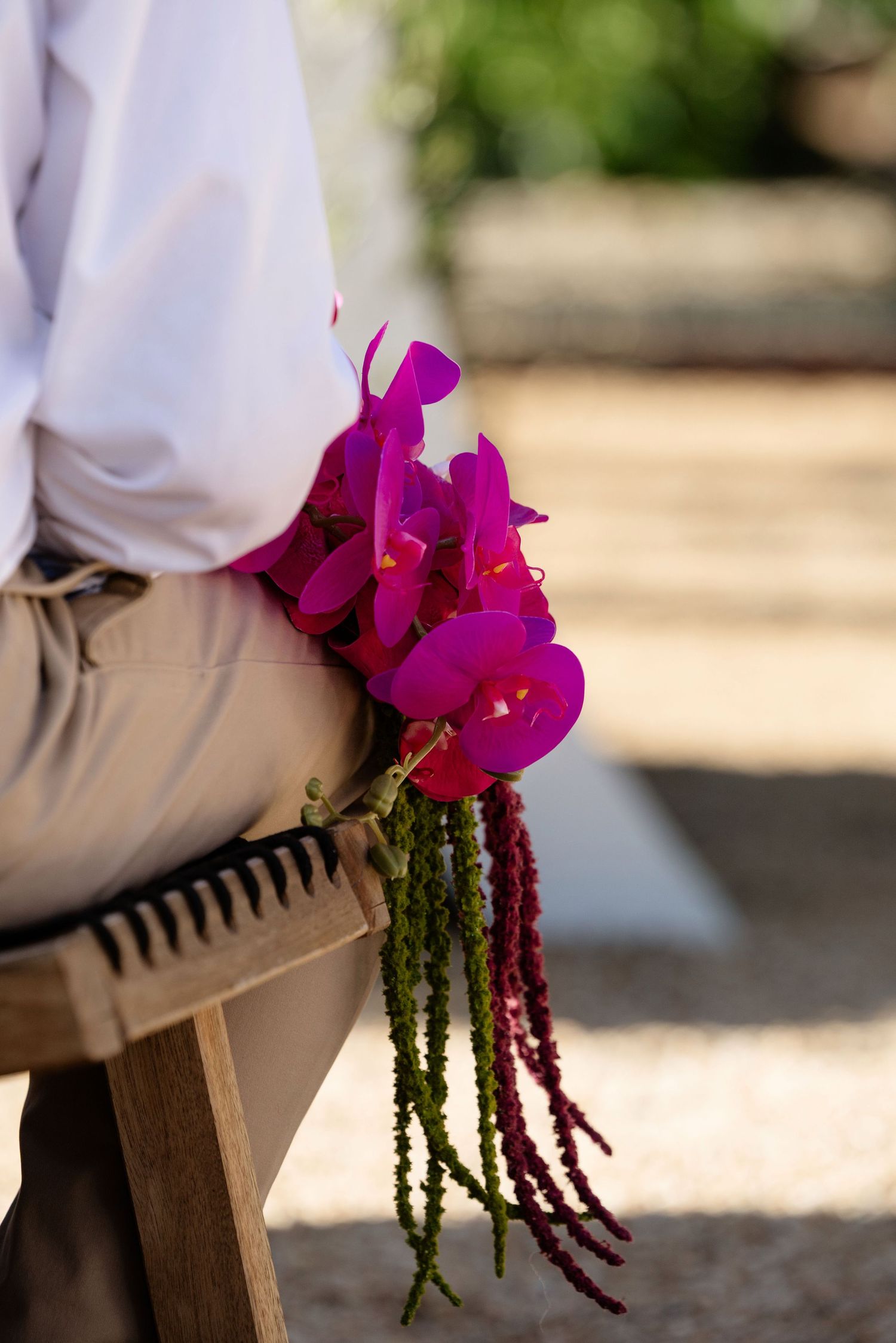 Close-up of vibrant magenta orchid and amaranth wedding bouquet resting on wooden chair at outdoor ceremony.