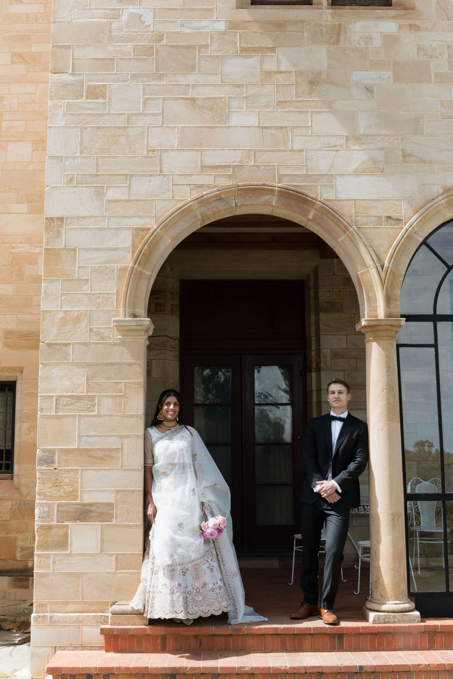 Bride in Indian white lehenga and groom in tuxedo standing under sandstone archway entrance.