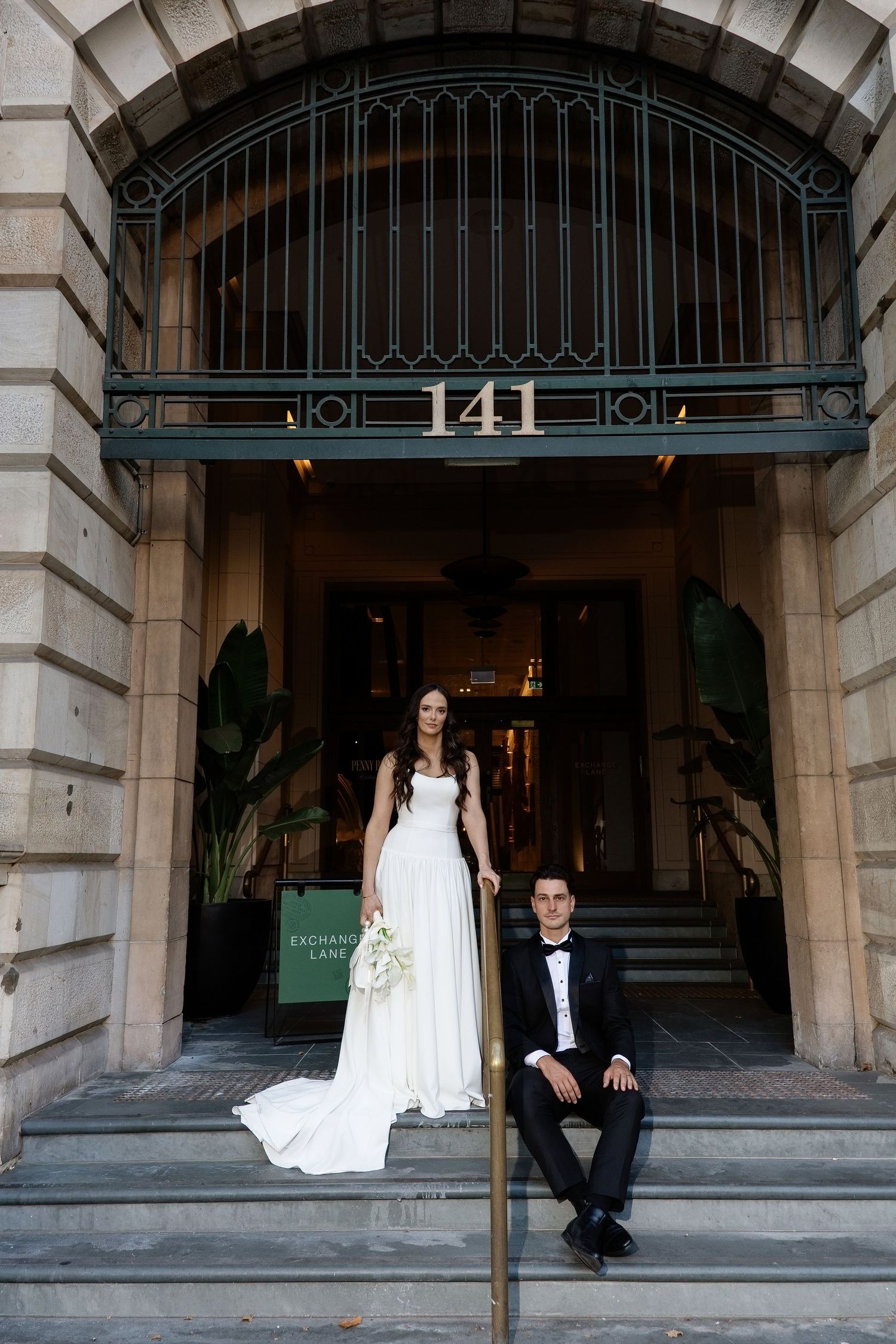 Bride standing and groom sitting on steps of a classic building entrance numbered 141.