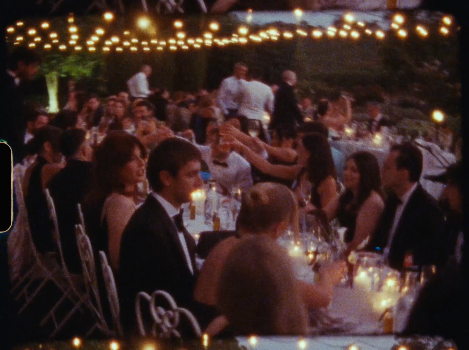 Guests celebrating at a candlelit outdoor wedding reception under warm string lights in the evening.