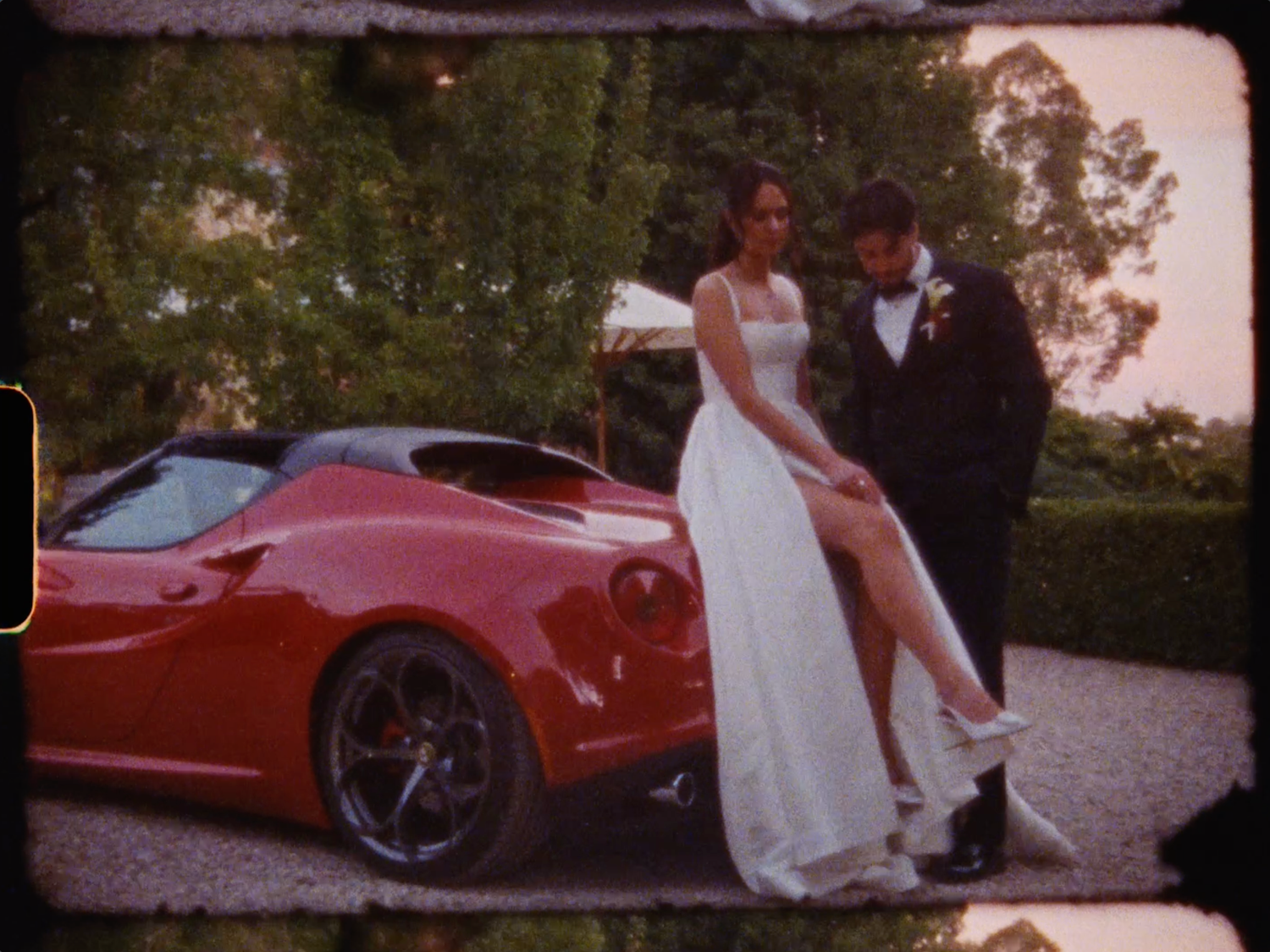 Bride in white slit gown posing with groom beside a red Ferrari sports car at sunset wedding.