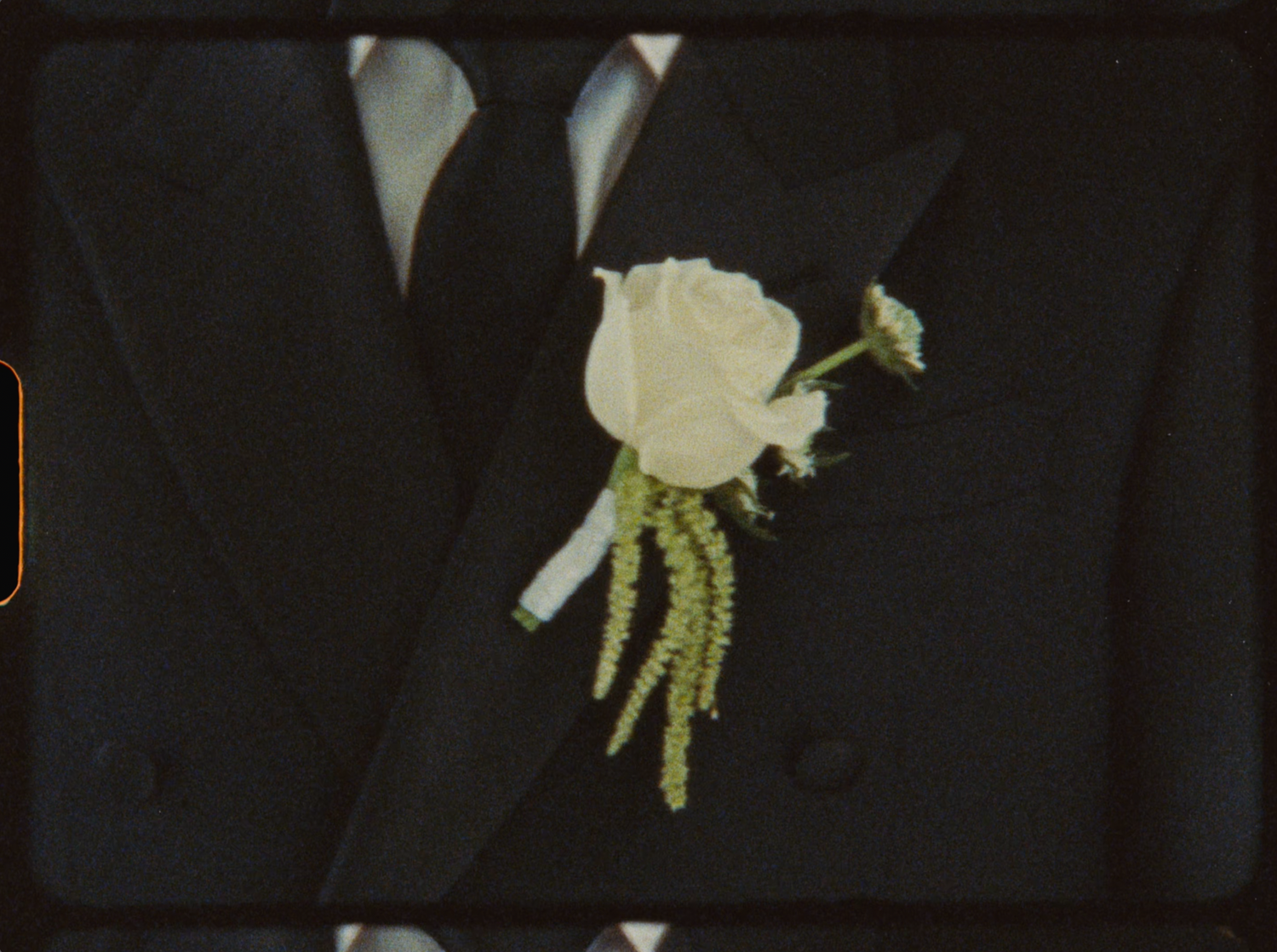 White rose boutonniere with green accents pinned to a black tuxedo lapel.
